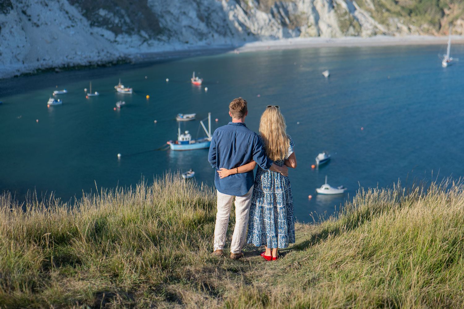 Couple standing on a grassy cliff overlooking a stunning turquoise bay with boats, embracing while enjoying the scenic coastal view.