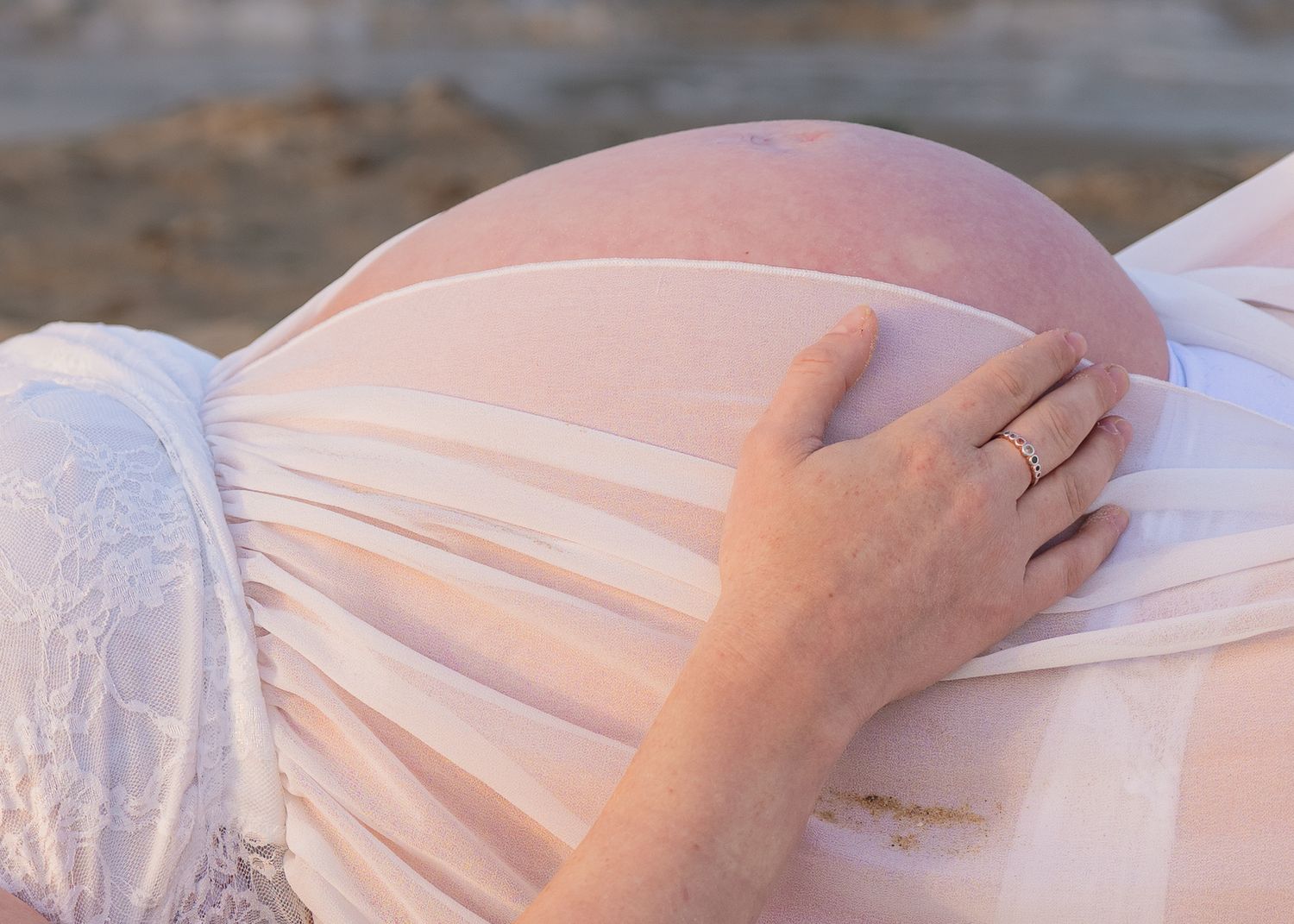Close-up of a pregnant woman's bare belly outdoors, with a hand wearing a ring gently resting on it.