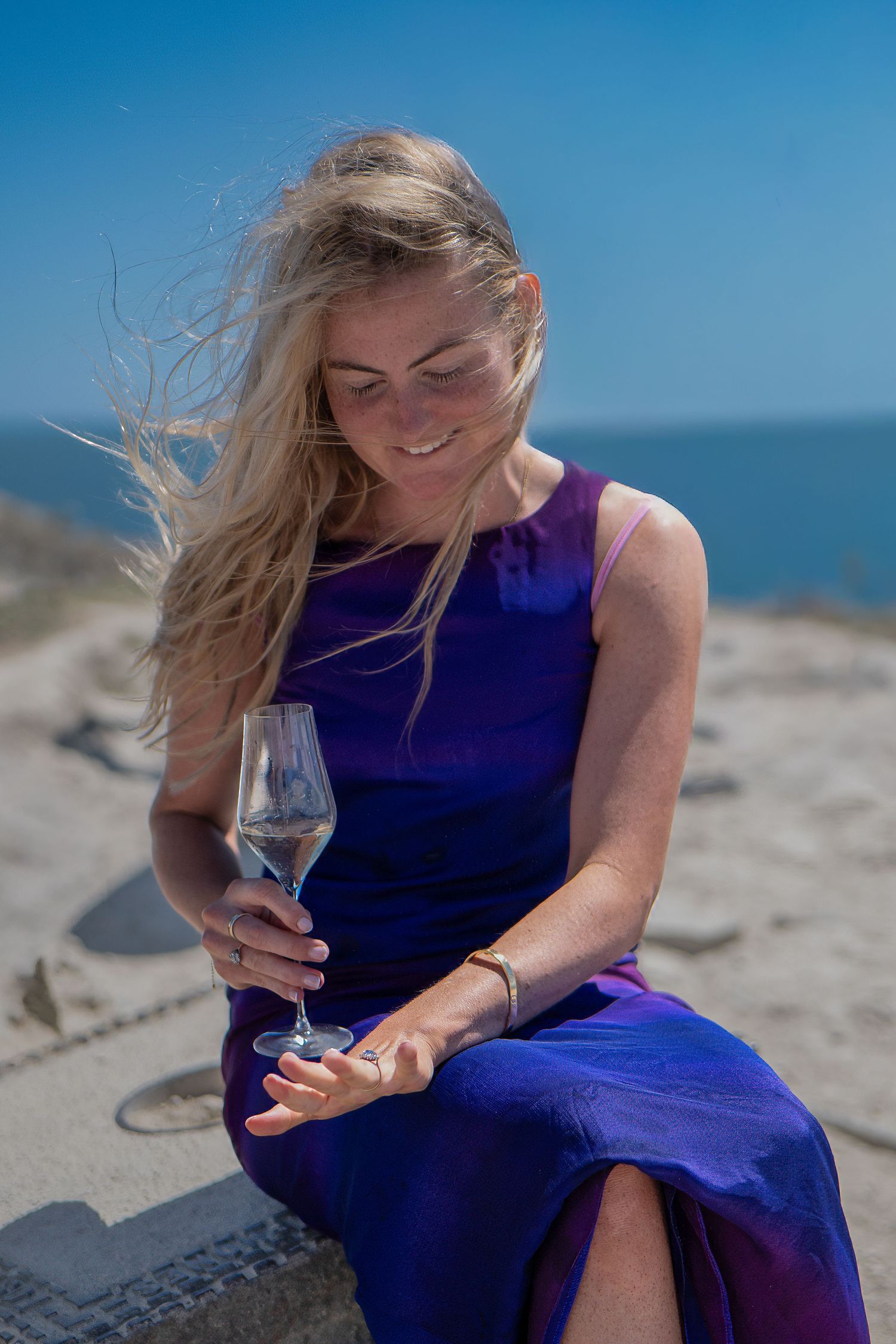 Smiling blonde woman in purple dress holding champagne glass on sandy beach with blue ocean background.
