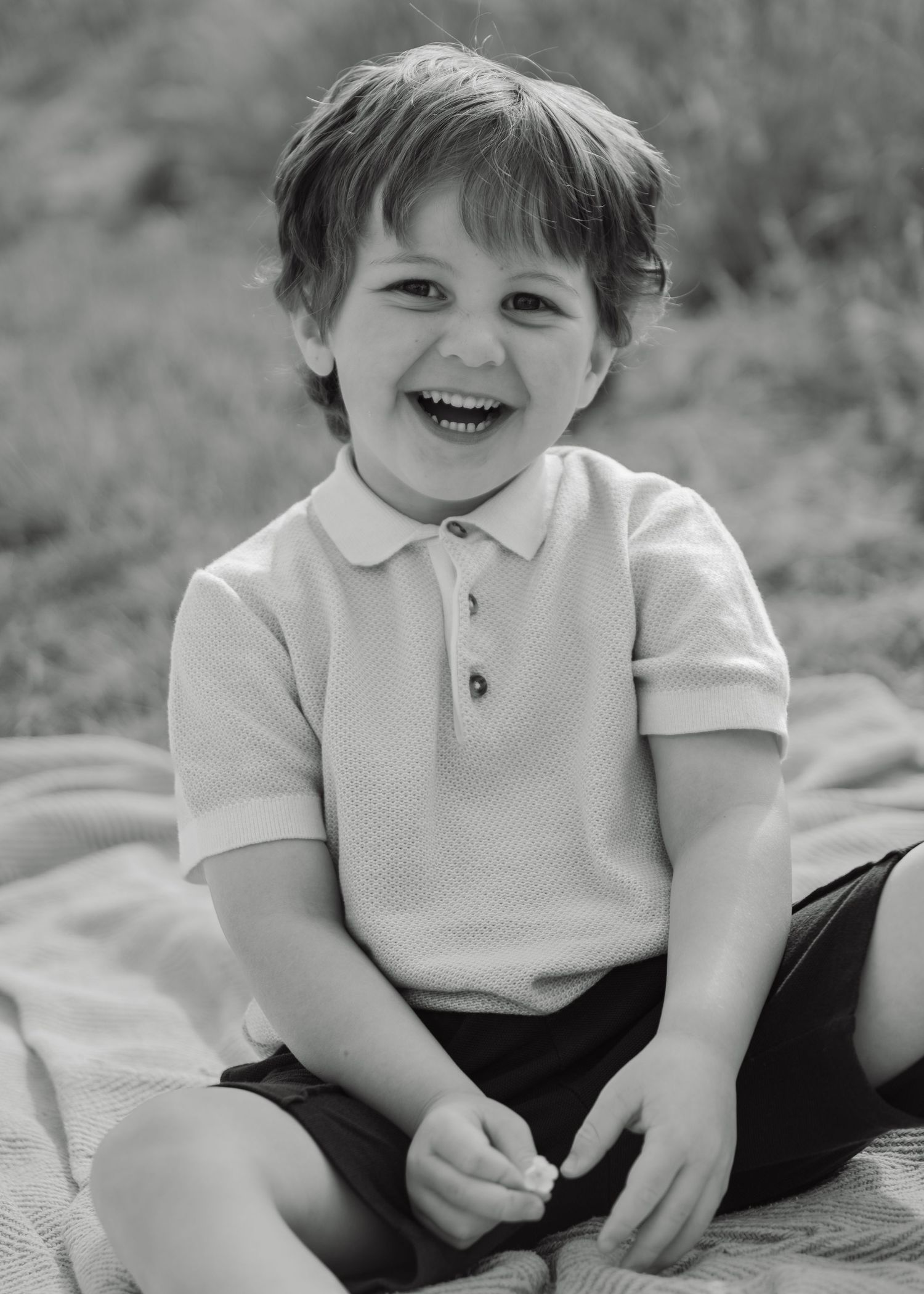 Black and white photo of a laughing young boy in a polo shirt sitting on a blanket outdoors.