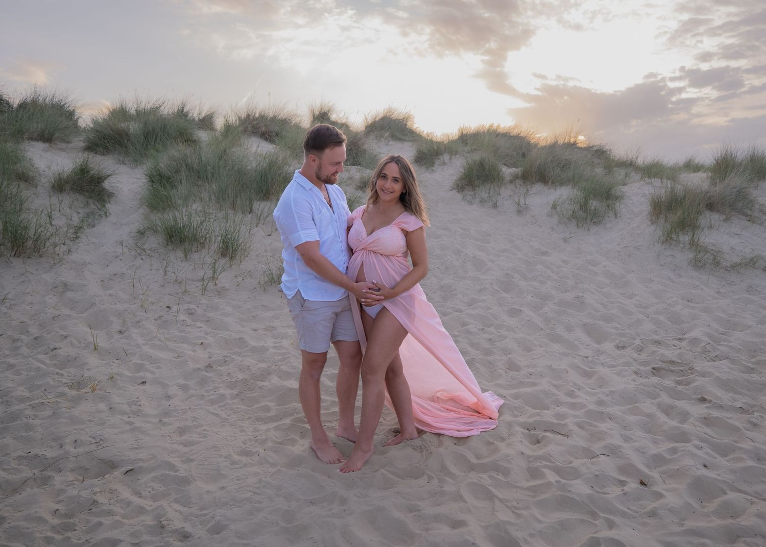 Couple poses on sandy beach at sunset, woman in pink maternity gown, man in white shirt, dunes in background.