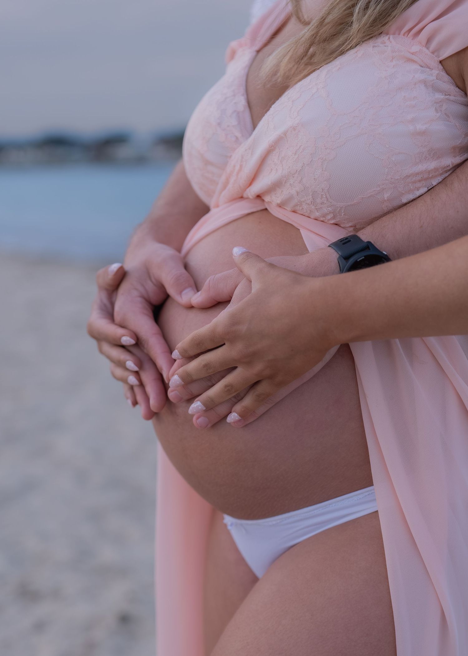 Pregnant woman in pink robe holding baby bump on beach, forming heart shape with hands.