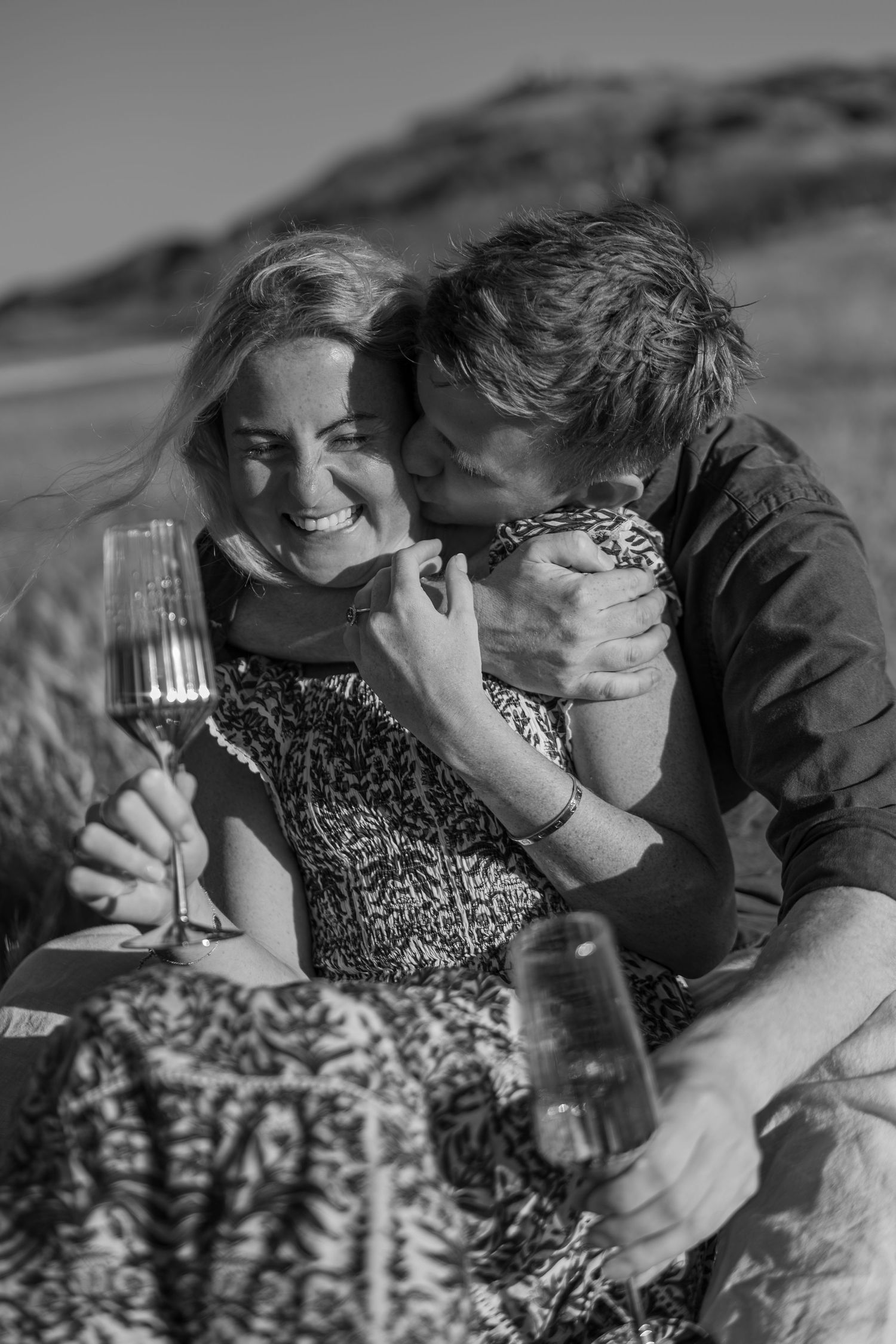 Happy couple sharing champagne on beach, man kissing womans cheek in black and white romantic photo.