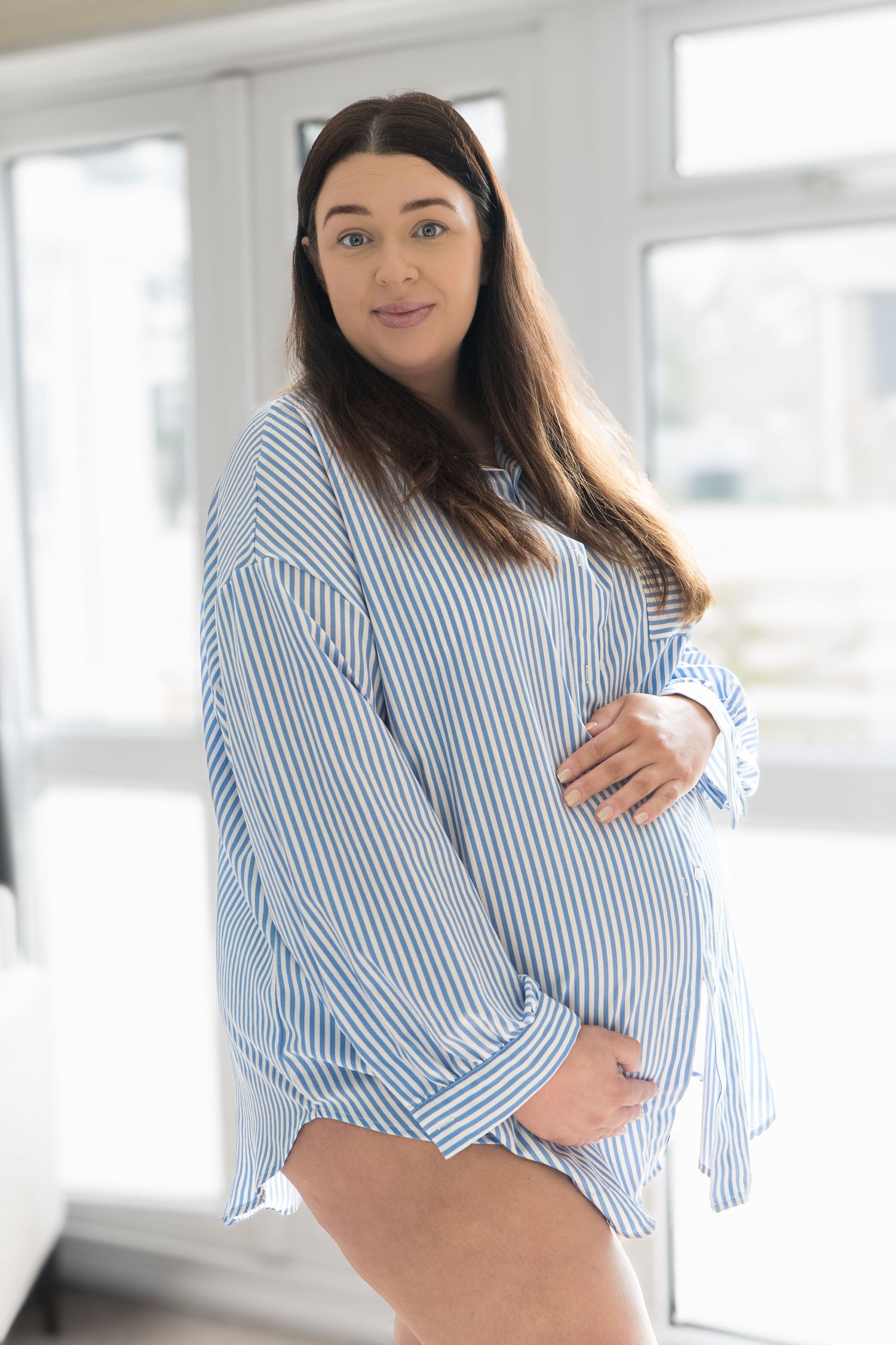 Pregnant woman in blue striped oversized shirt cradling baby bump, standing indoors with natural light.