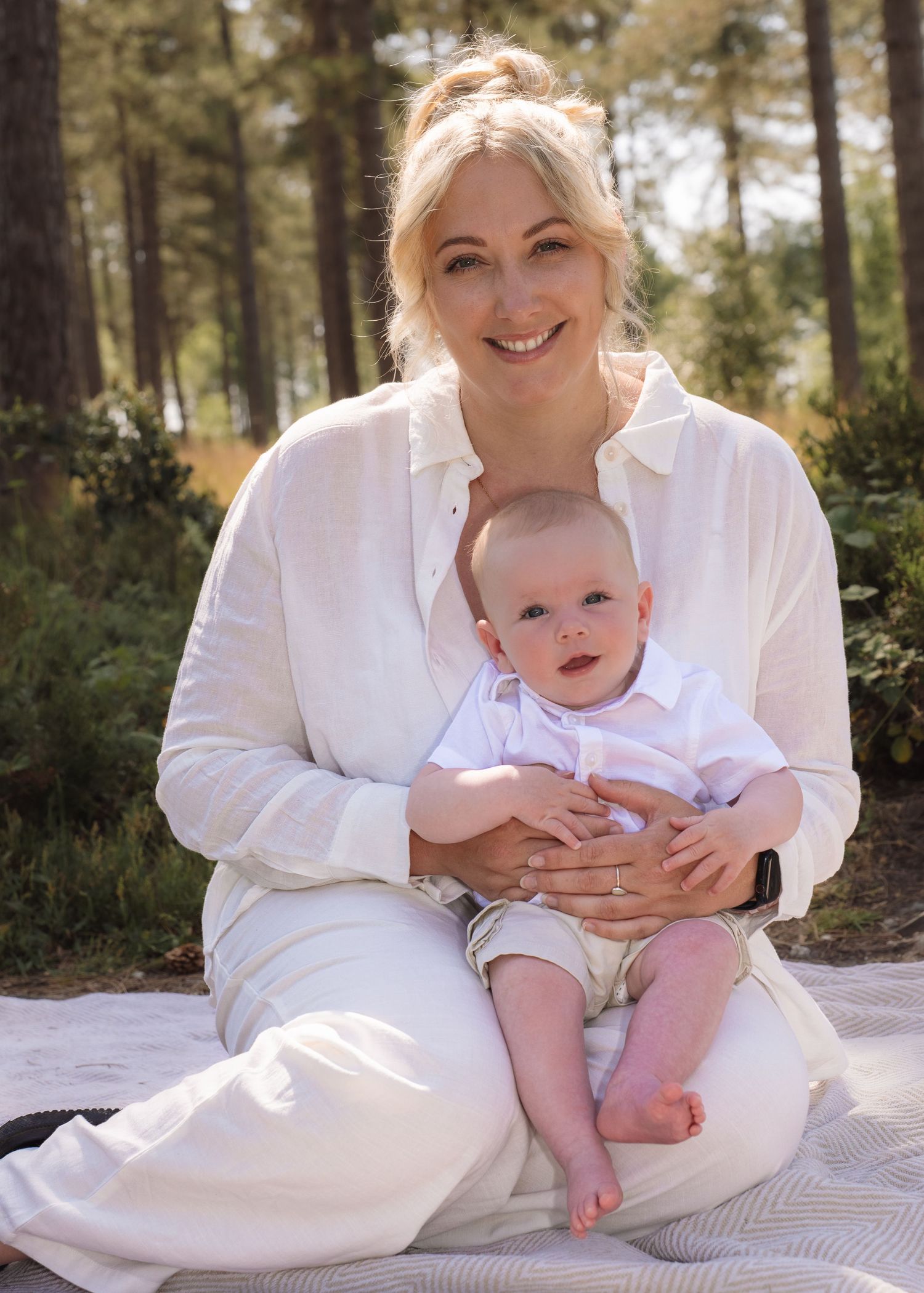 Smiling blonde woman in white linen shirt sitting outdoors holding a cute baby boy on her lap.