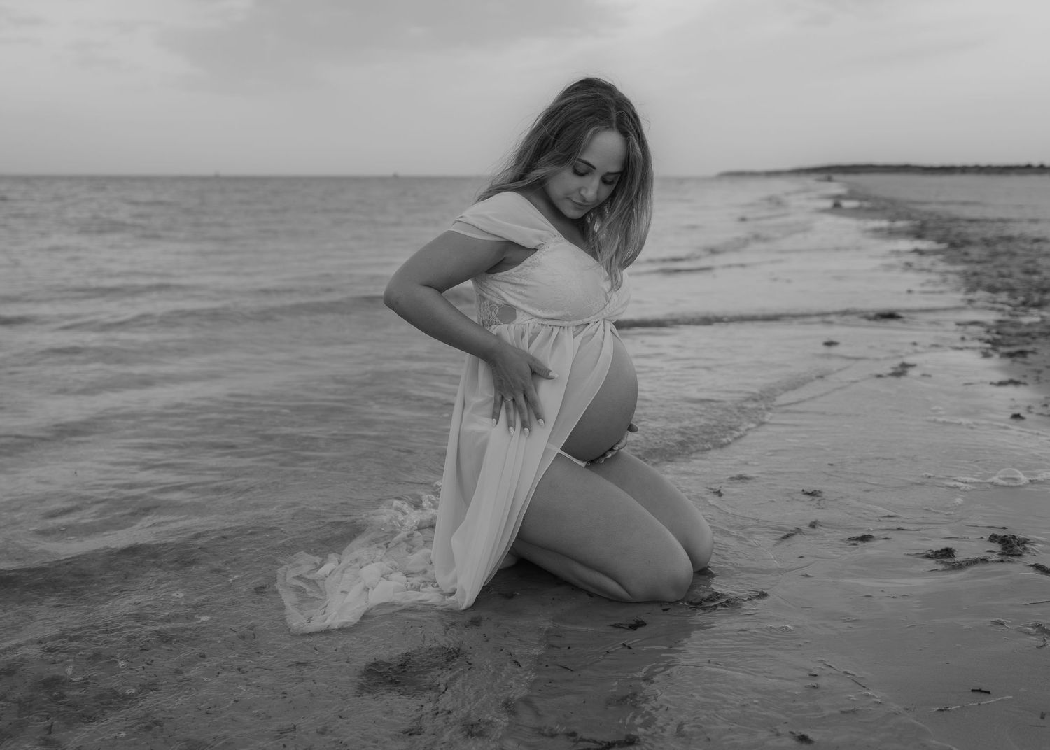Black and white maternity photo of pregnant woman in white dress kneeling on beach shore with ocean waves.