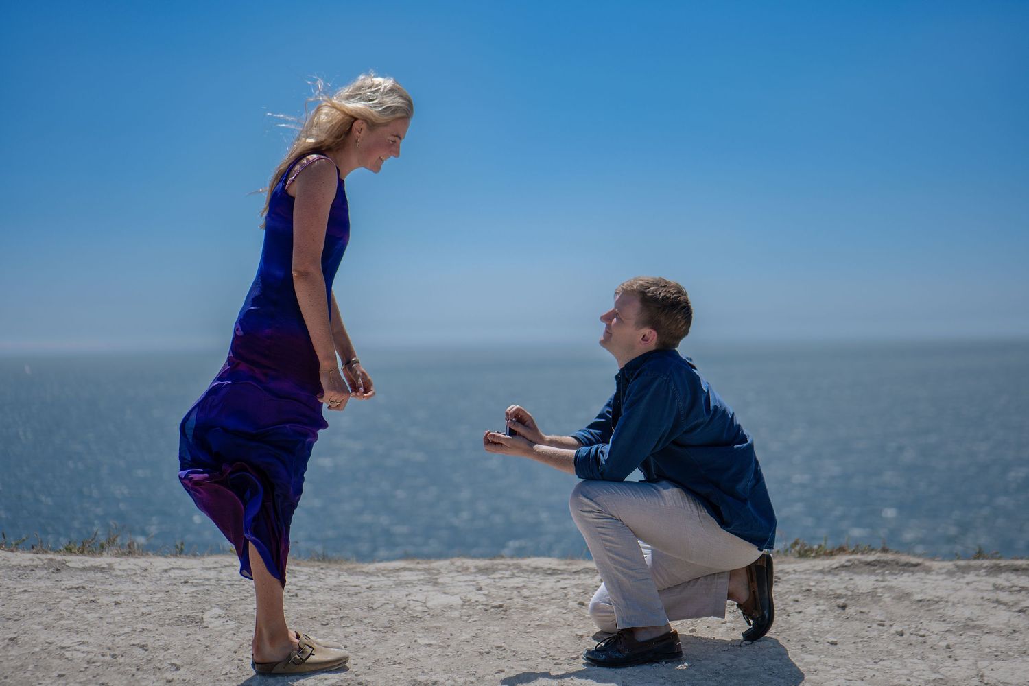 Man kneeling on one knee proposing to a woman in a blue dress by the ocean on a sunny day.