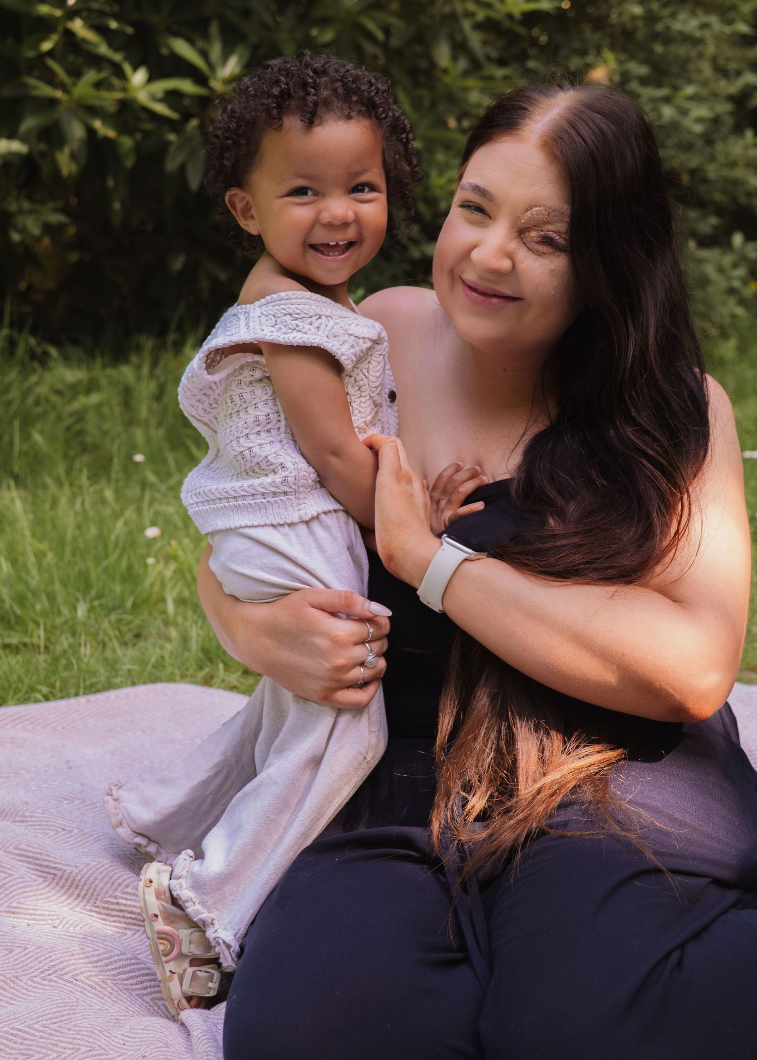 A smiling woman with long dark hair holds a happy curly-haired toddler outdoors on a sunny day.