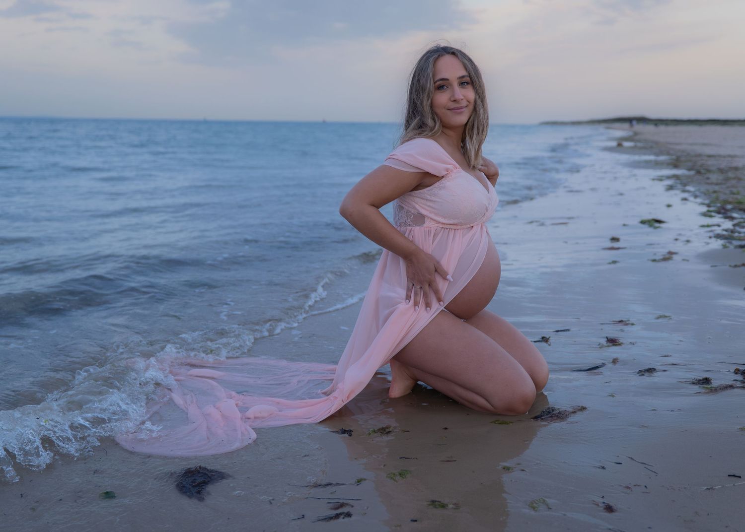 Pregnant woman in flowing pink gown posing on sandy beach at dusk with ocean waves in background.