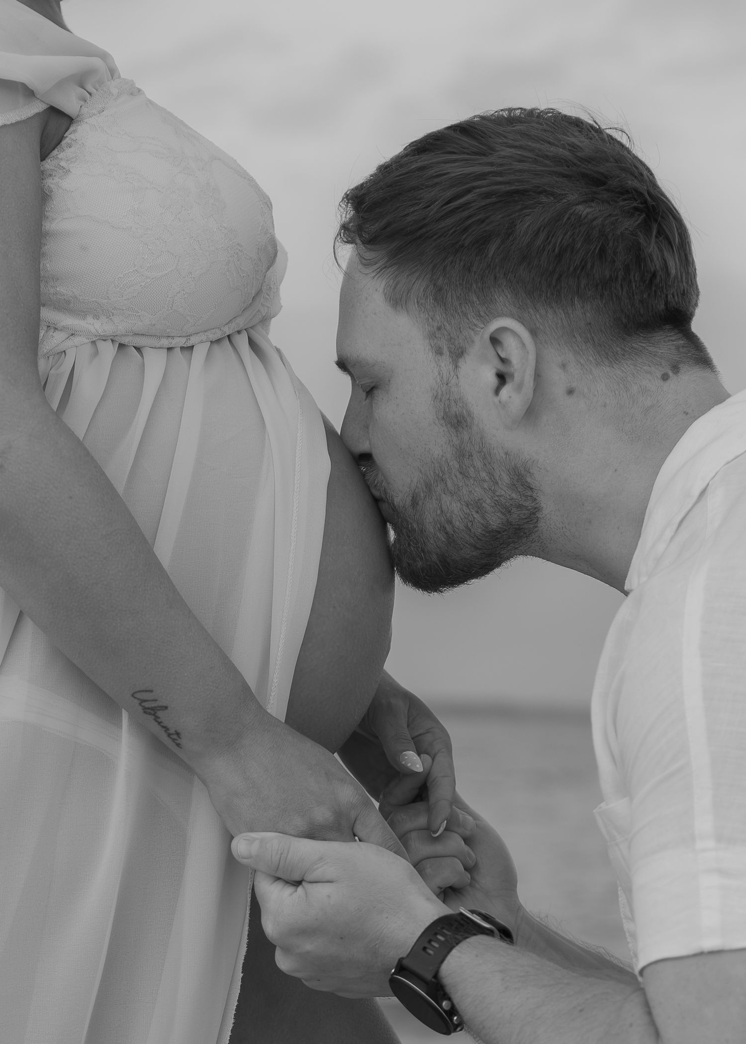 A man tenderly kisses a pregnant woman's belly in a black and white maternity photo shoot outdoors.