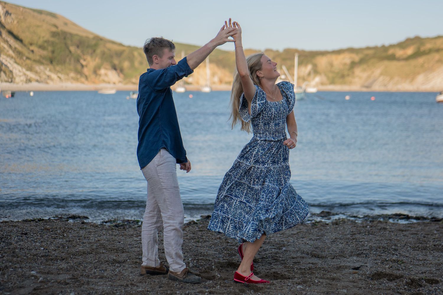Couple dancing together on a pebble beach with scenic coastal cliffs and calm blue sea in background.