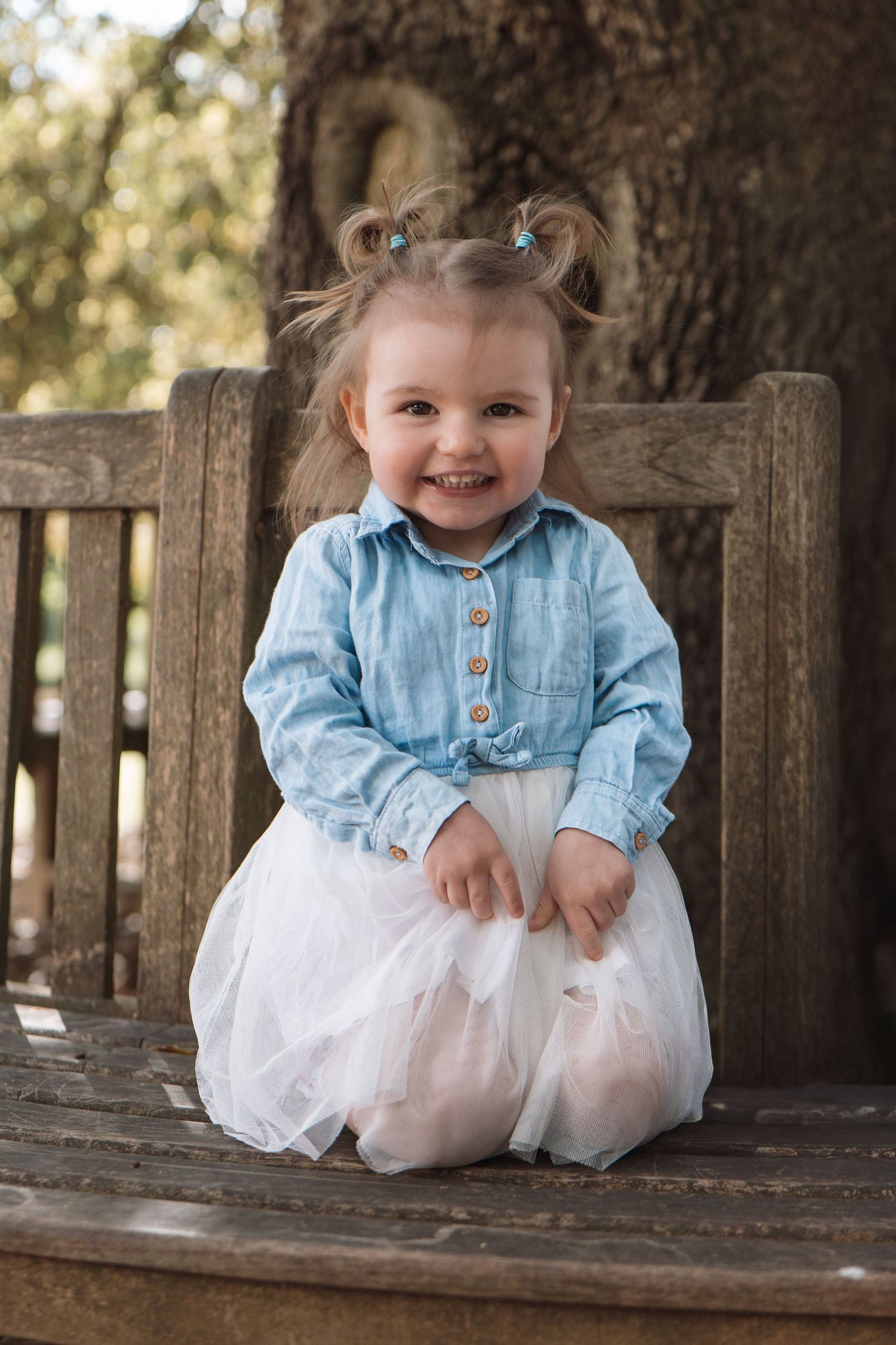 Happy little girl in blue denim shirt and white tulle skirt smiling on a wooden bench.