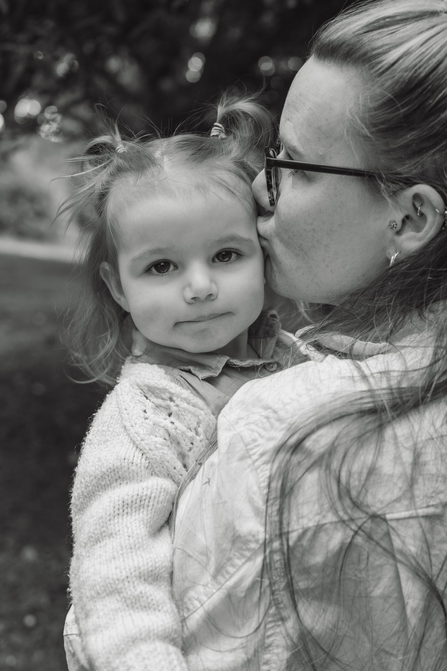 A mother with glasses tenderly kisses her young toddler daughter on the cheek in black and white.