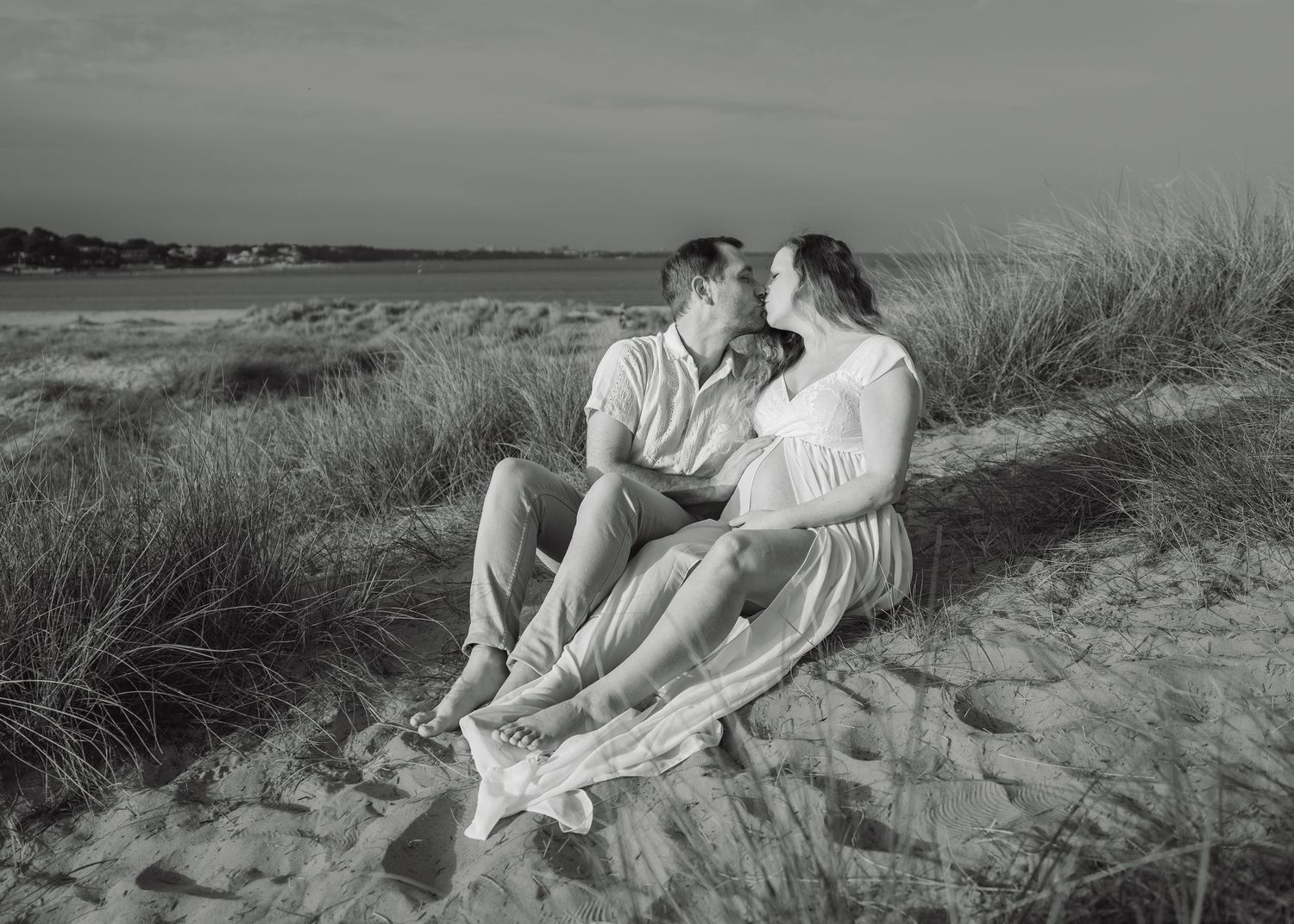Black and white photo of a couple sitting on sand dunes, the woman visibly pregnant, sharing a tender moment by the beach.