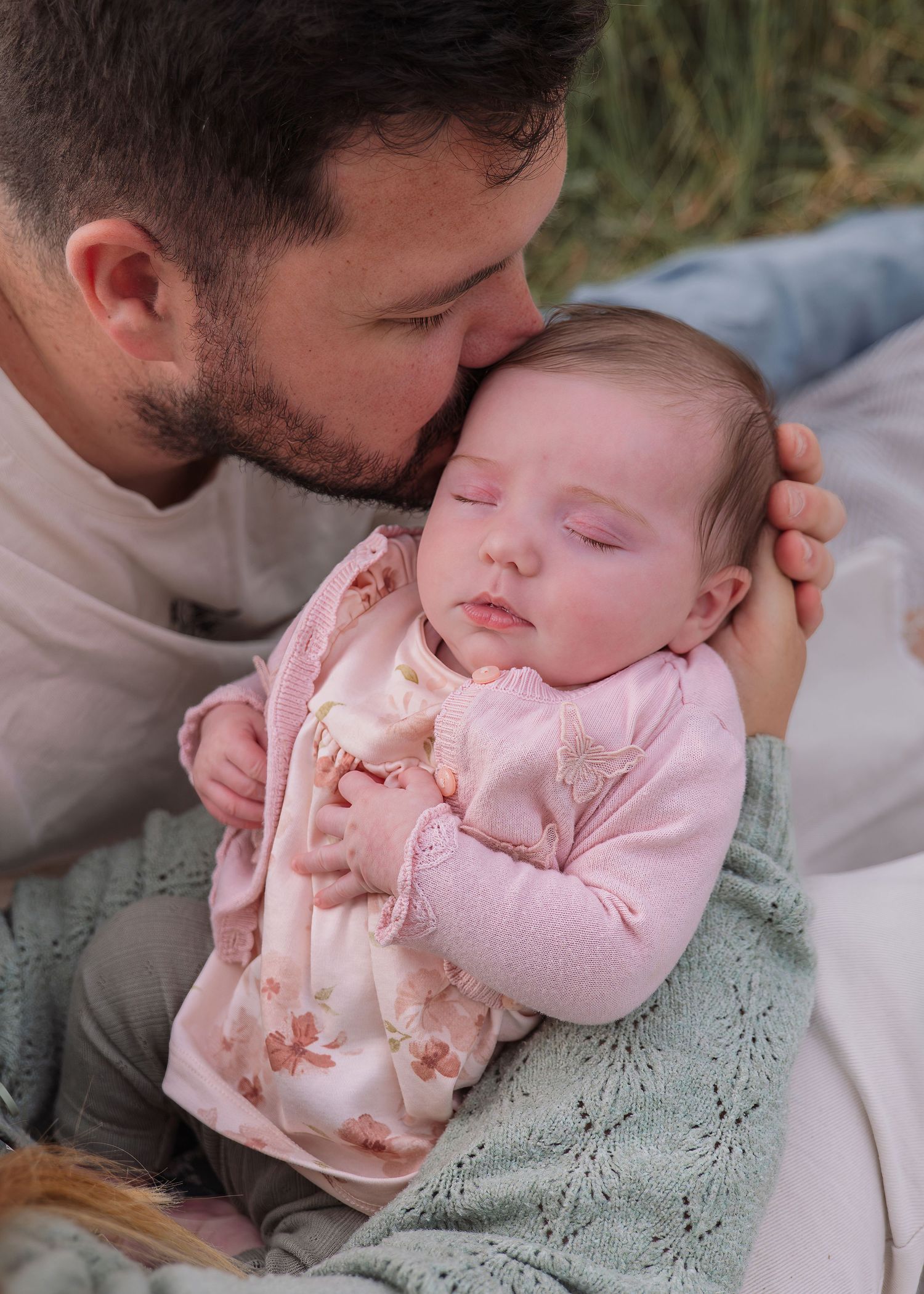 A father tenderly kisses his sleeping newborn baby dressed in a pink floral outfit while cradling her gently.