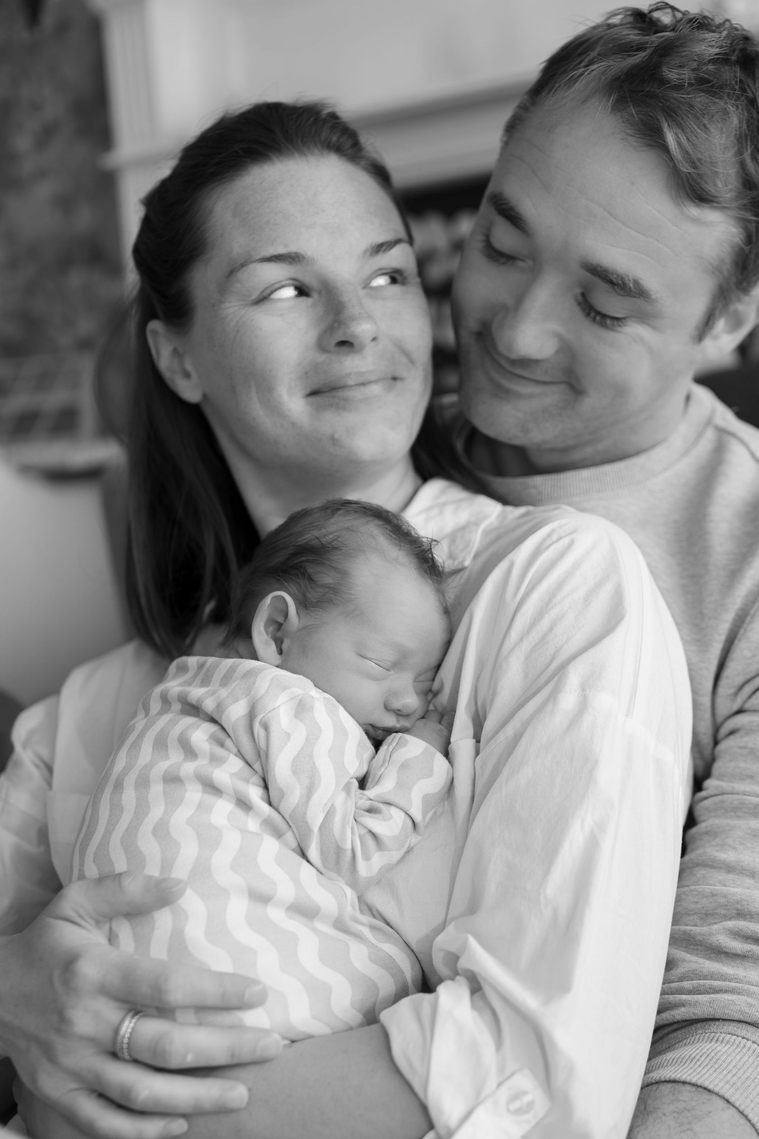 Black and white photo of smiling new parents holding their sleeping swaddled newborn baby.