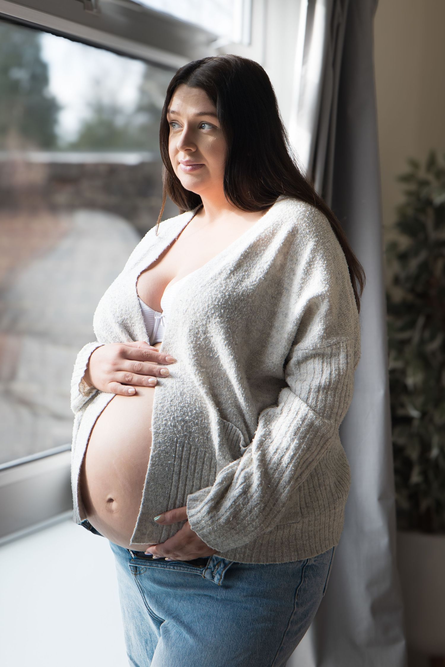 Pregnant woman in grey cardigan and jeans cradling baby bump while gazing out a window.