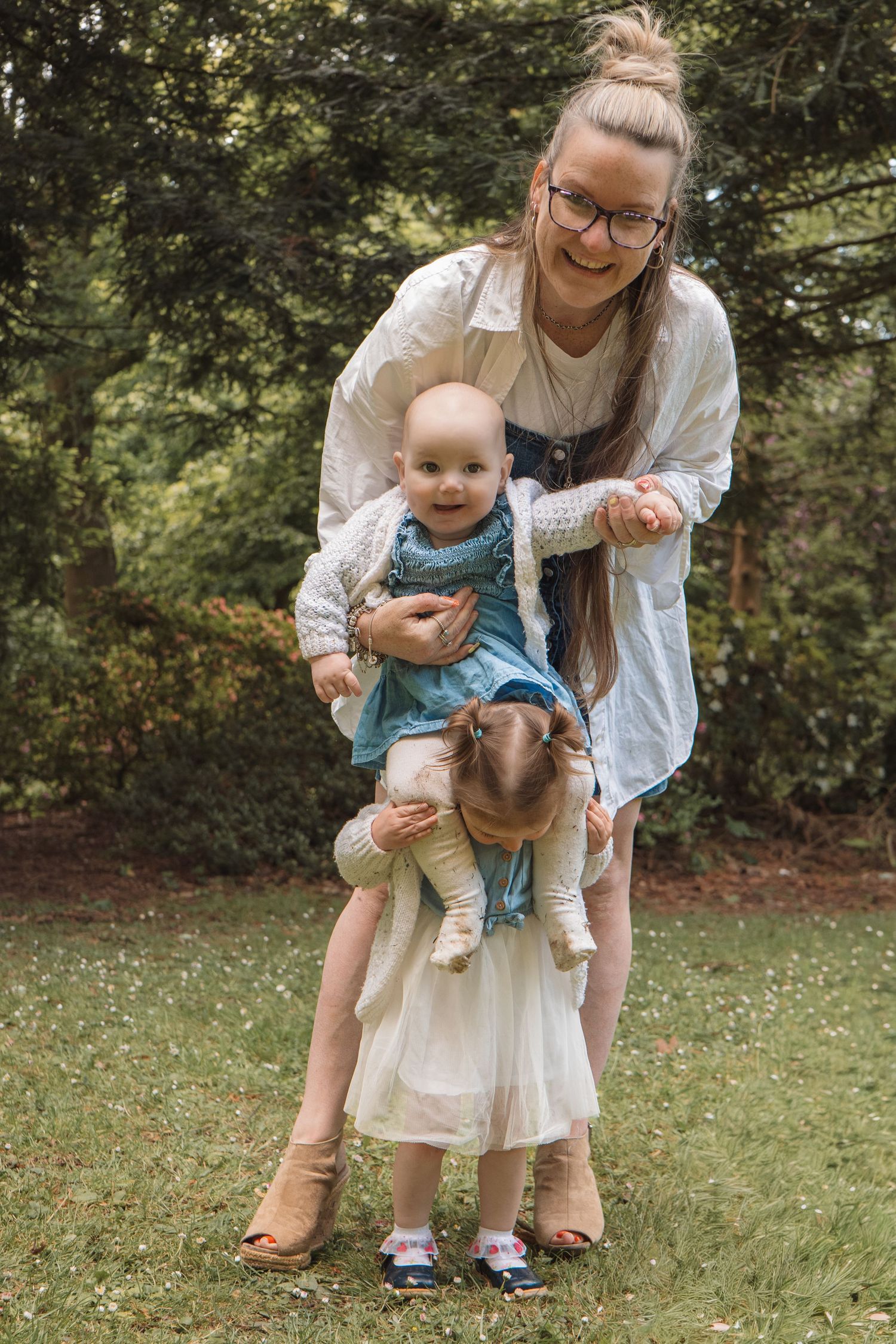 A smiling mother with glasses holds a baby while a toddler girl plays outdoors in a lush green park.