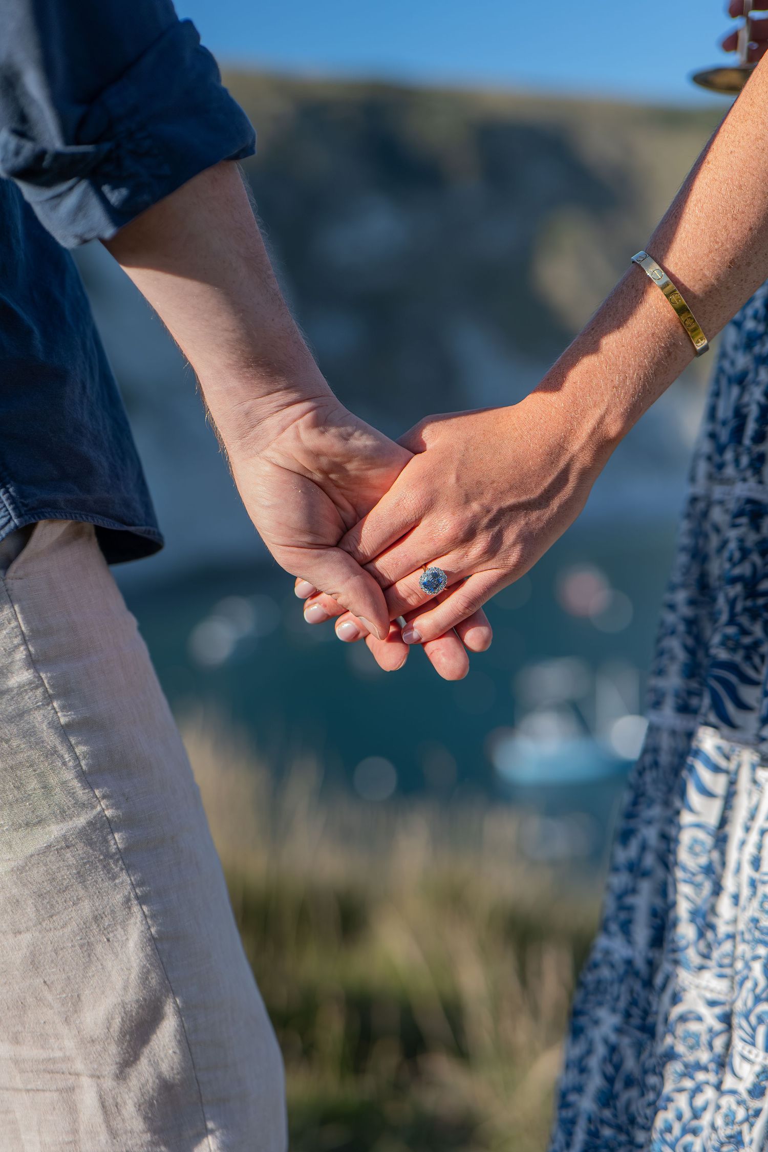 Couple holding hands outdoors with a turquoise ocean and cliffs in the background, woman wearing a blue gemstone ring.