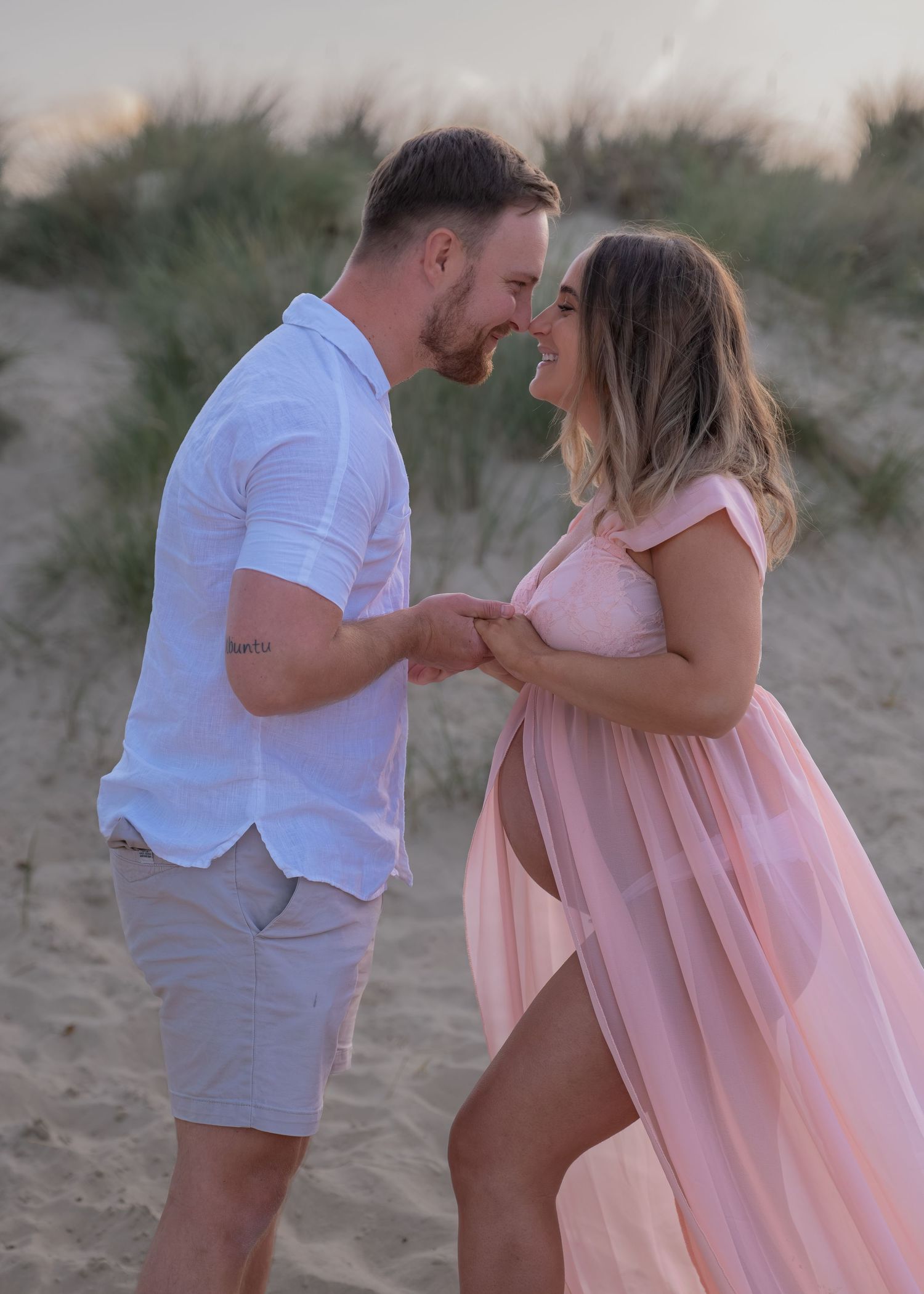 Couple sharing a romantic moment on a sandy beach, woman in pink maternity gown holding hands with man in white shirt.