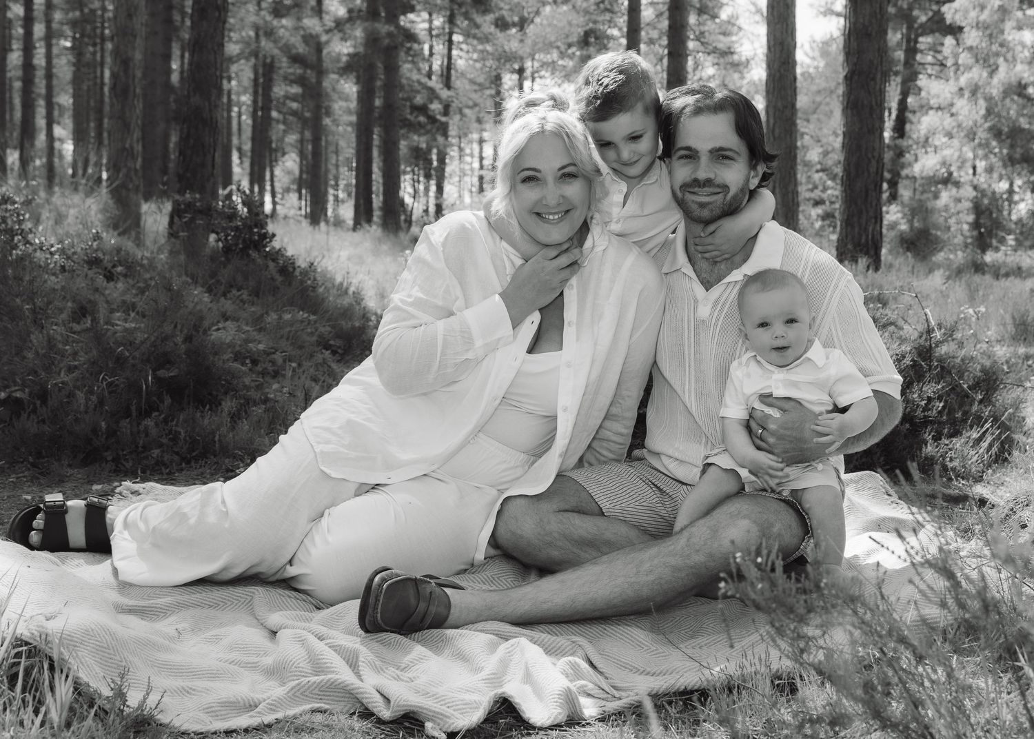 Black and white family photo of parents and two young children sitting on a blanket in a pine forest.