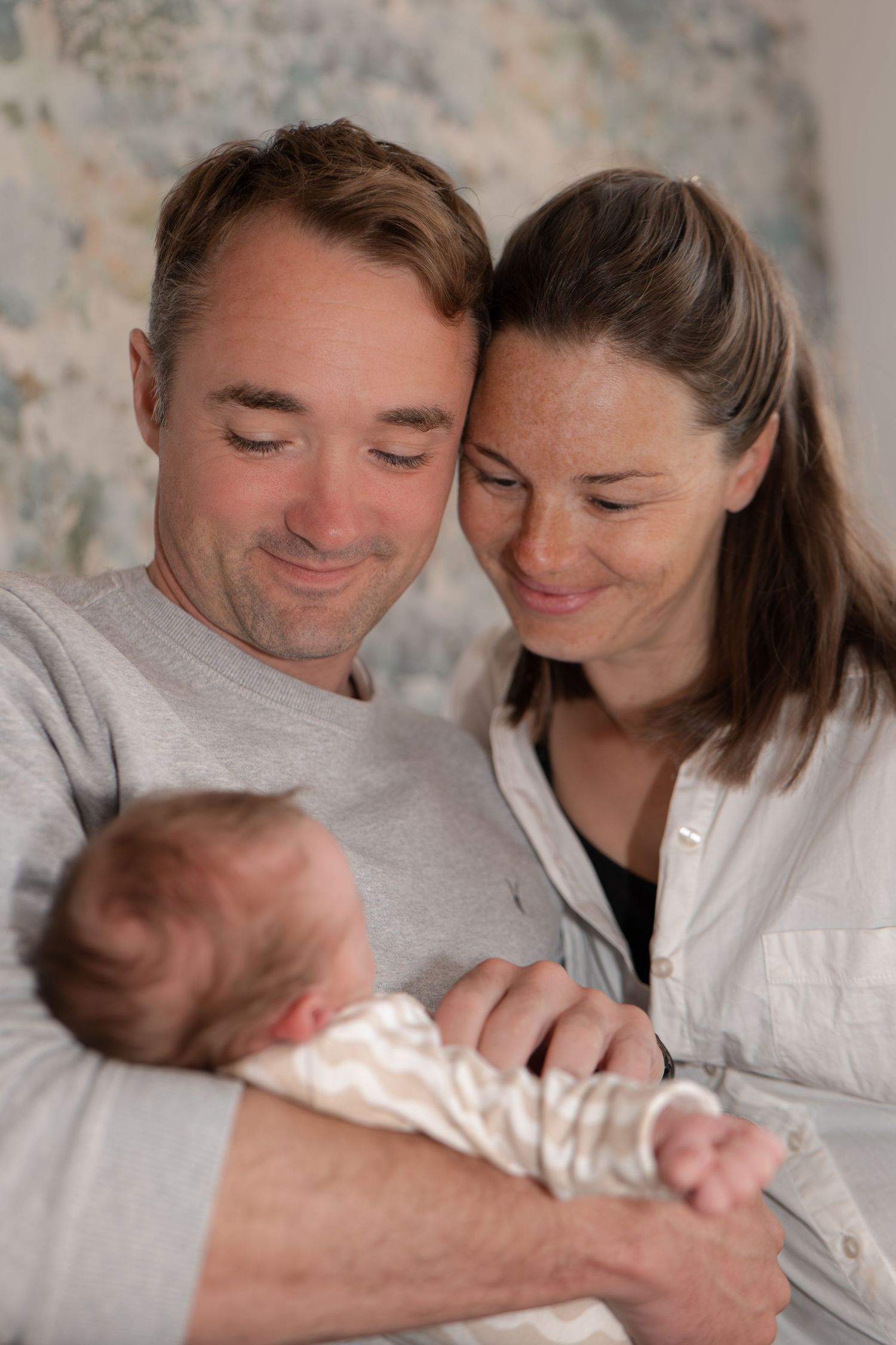A couple smiles lovingly at their newborn baby, with heads touching, in a warm and tender family portrait.