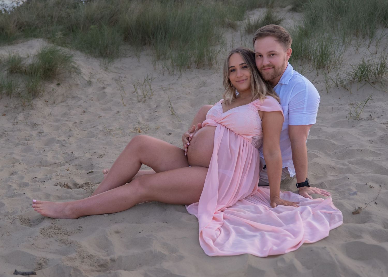Pregnant couple posing on sandy beach, woman in pink maternity dress, man in blue shirt, maternity photoshoot.