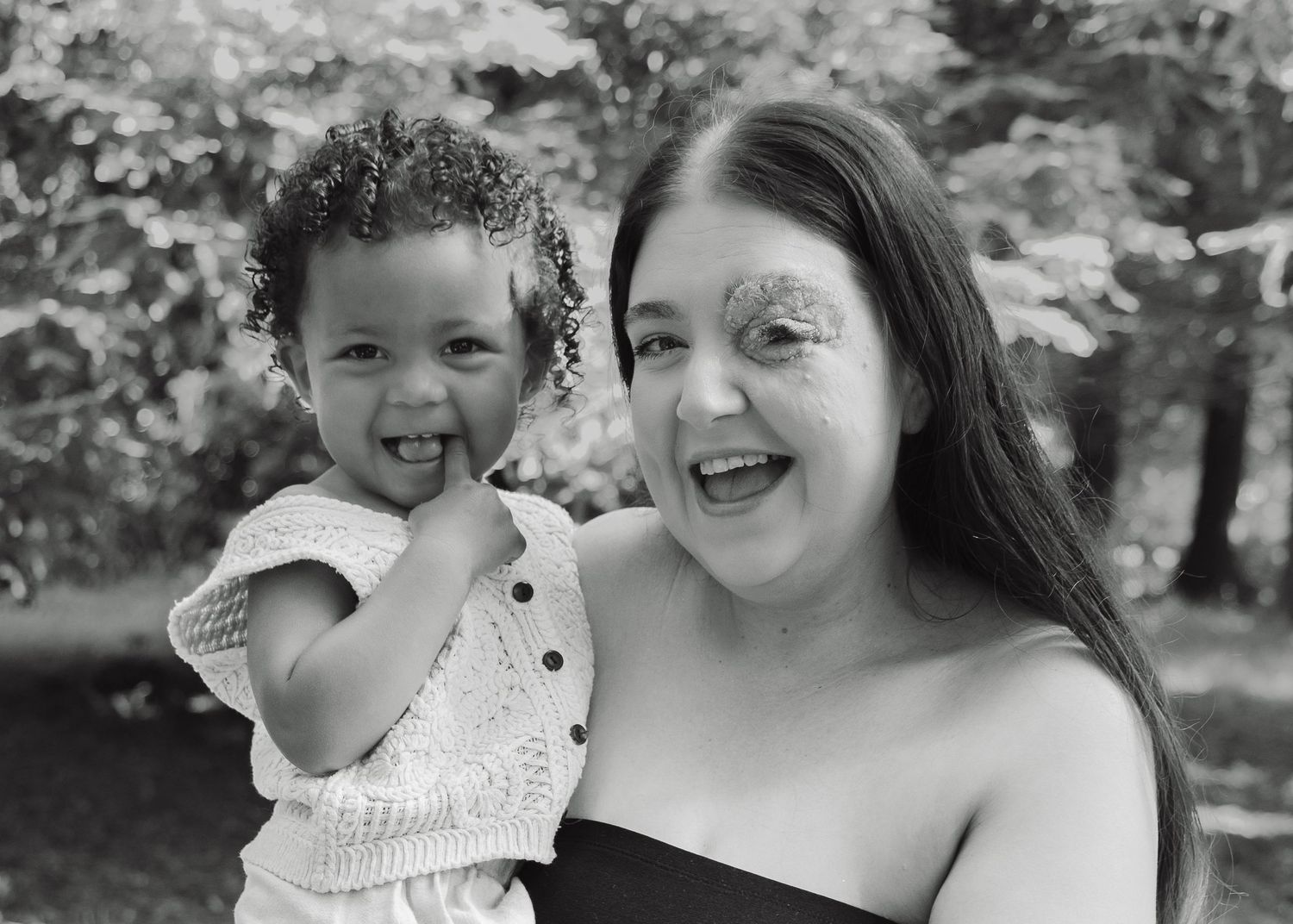 Black and white photo of a smiling woman and happy toddler girl posing together outdoors among trees.