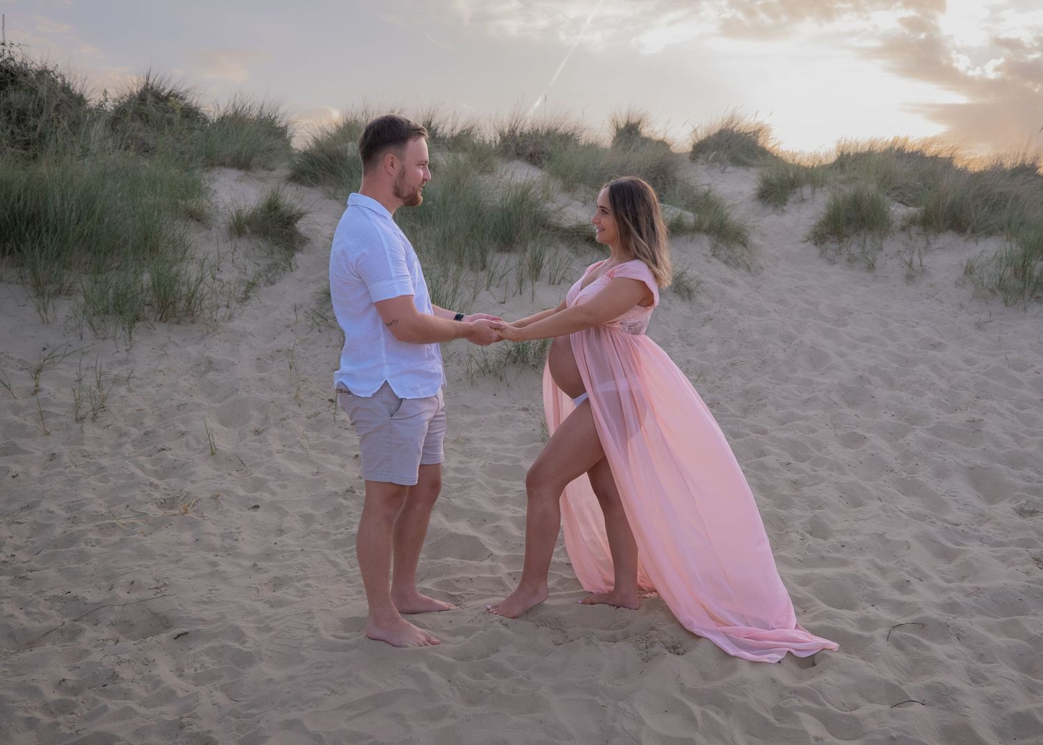 Couple holding hands on sandy beach at sunset, woman in flowing pink maternity gown, man in white shirt and gray shorts.