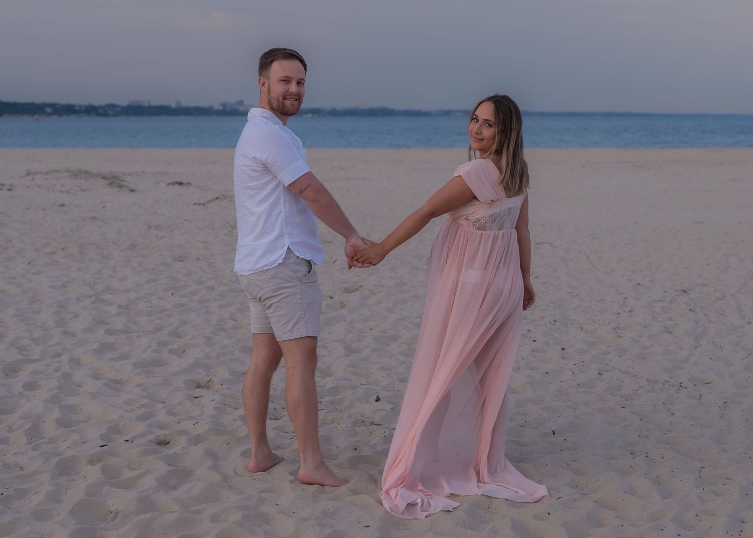 Couple holding hands on sandy beach at sunset, woman in flowing pink maternity gown, man in white shirt and shorts.