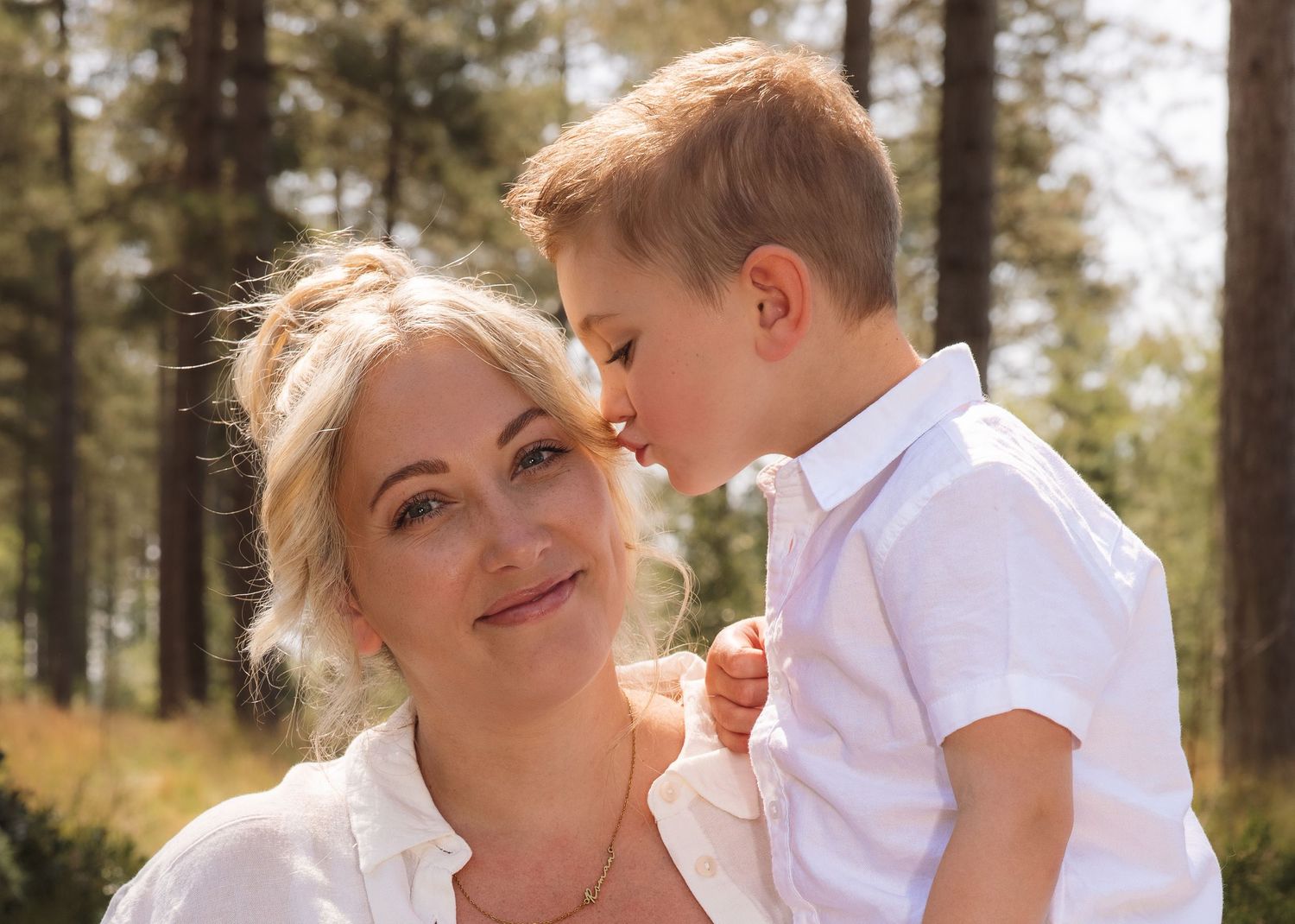 A blonde woman in a white shirt holds a young boy who gently kisses her cheek outdoors among trees.
