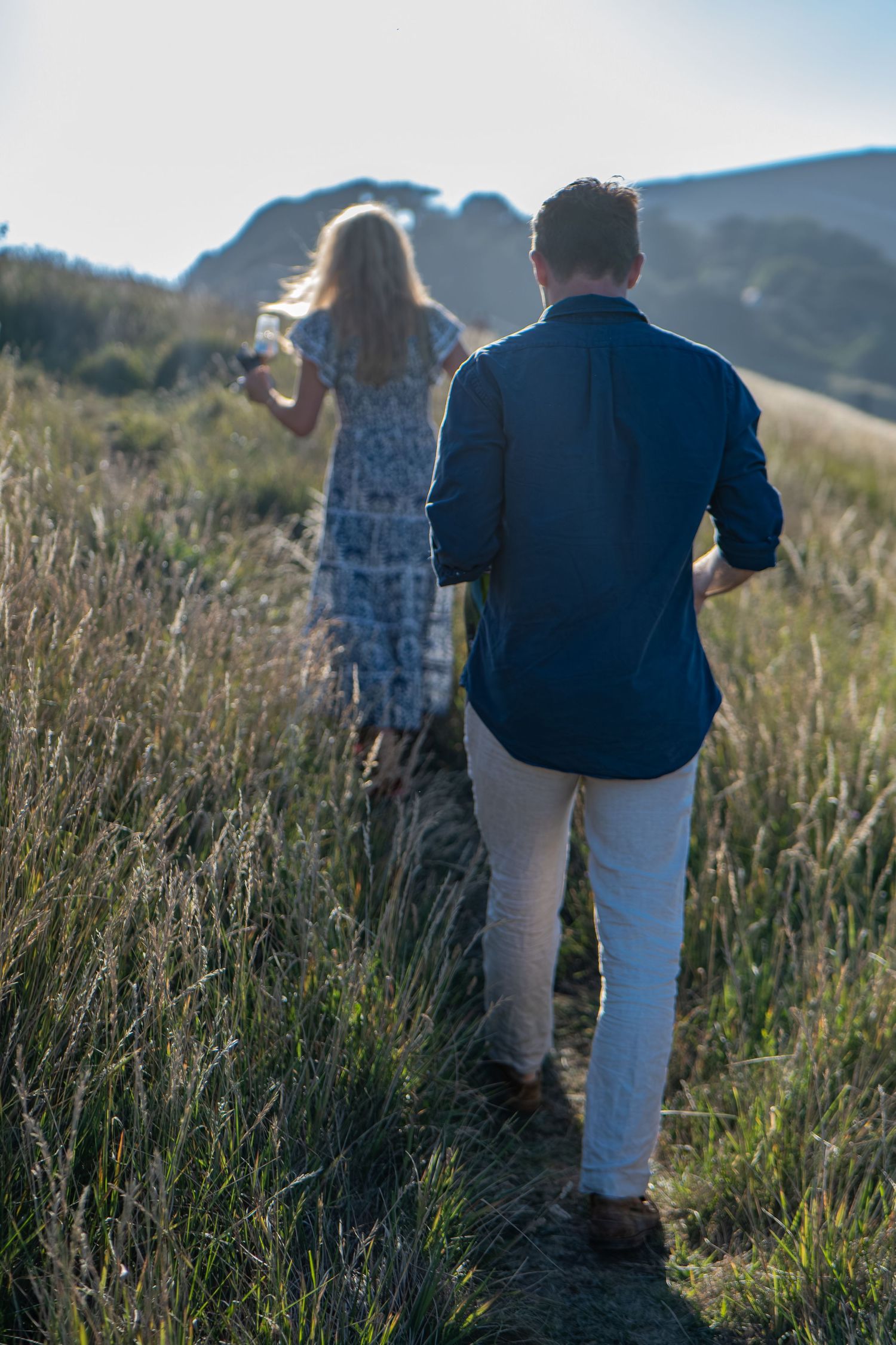 Couple walking through golden grass fields toward mountains, woman in floral dress, man in navy shirt holding wine glasses.