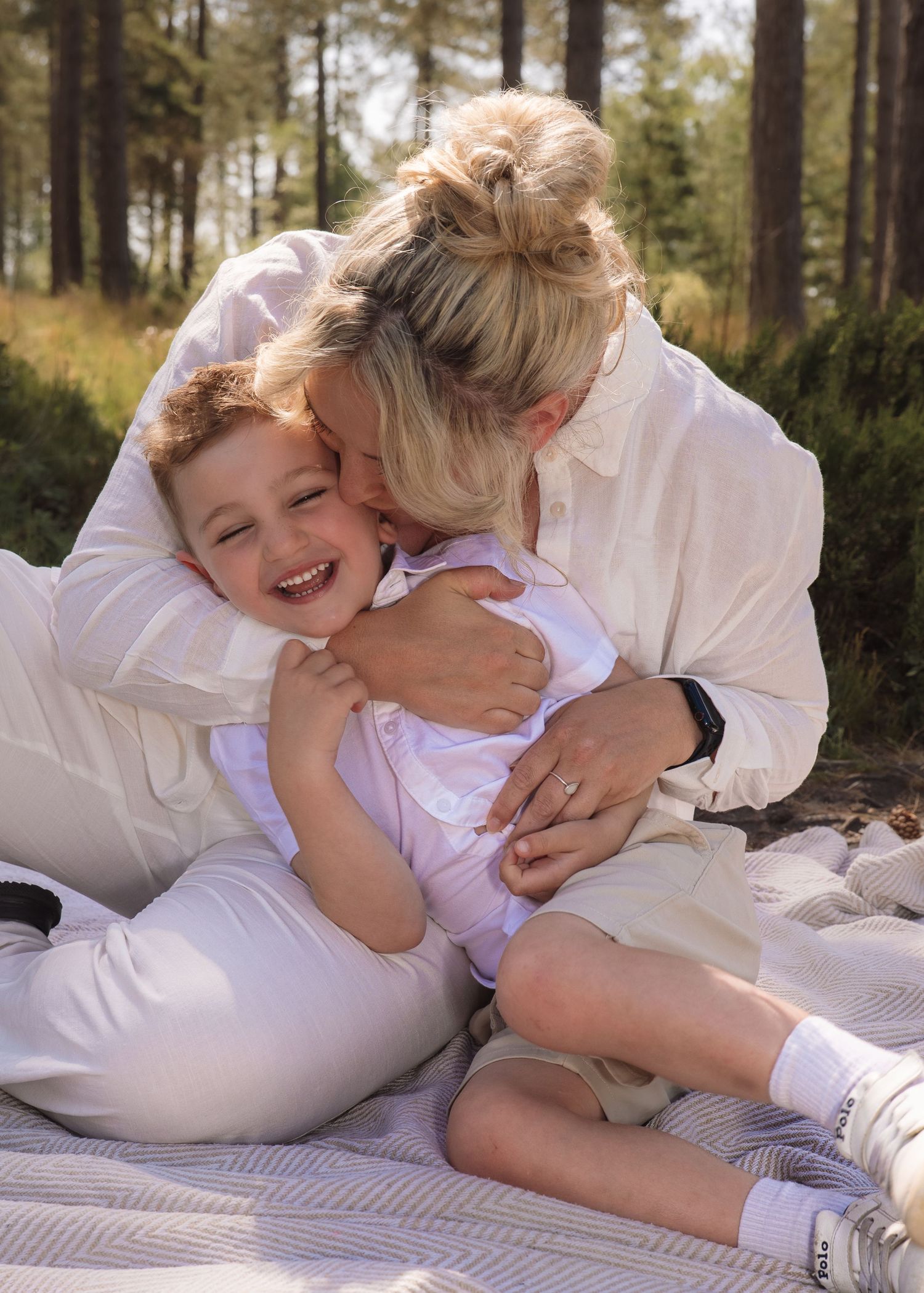 A joyful mother hugging and kissing her laughing young son while sitting on a blanket in a sunny forest.