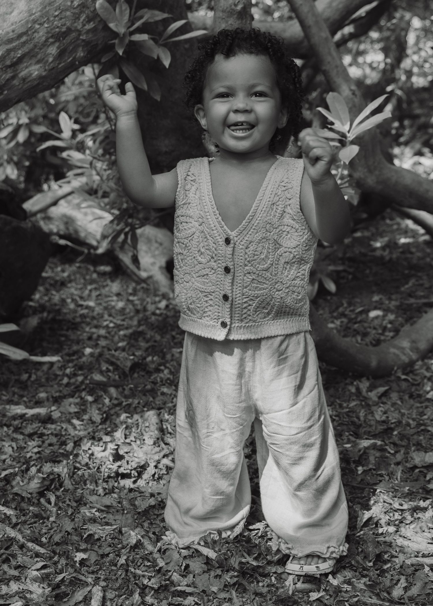 A smiling toddler in a knit vest and wide-leg pants stands outdoors among trees in a black and white photo.