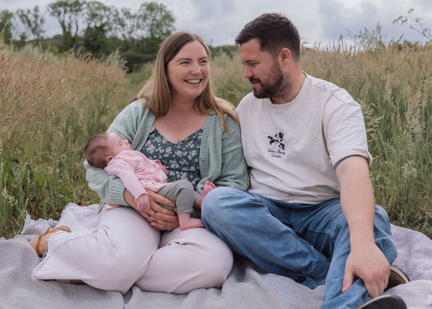 A happy couple sits in a meadow holding their newborn baby, smiling at each other on a sunny day.
