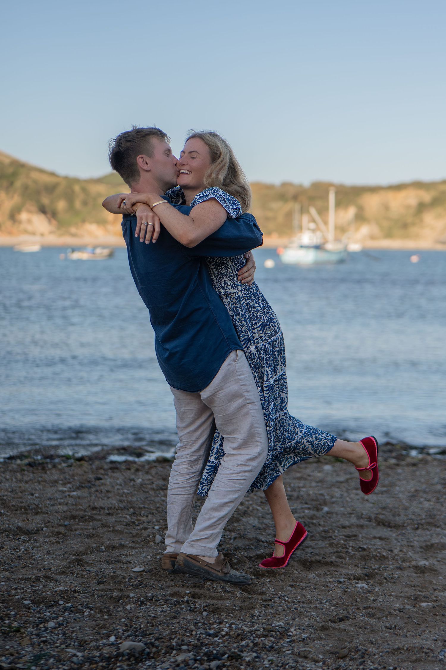 Couple embracing and kissing on a rocky beach at dusk, with sailboats and coastal cliffs in the background.