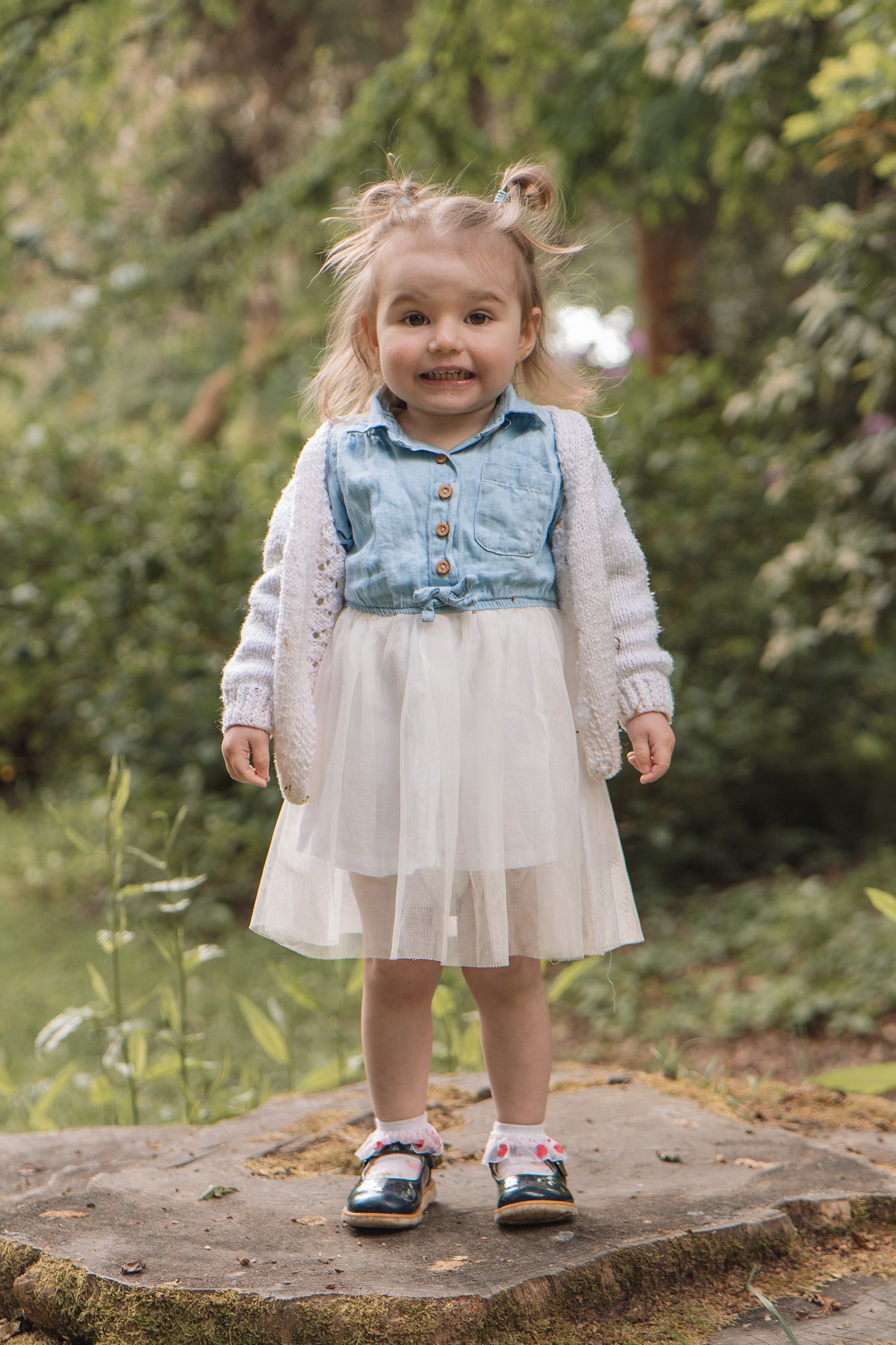 A cheerful toddler girl in a blue denim top and white tulle skirt stands on a stone in a green park.