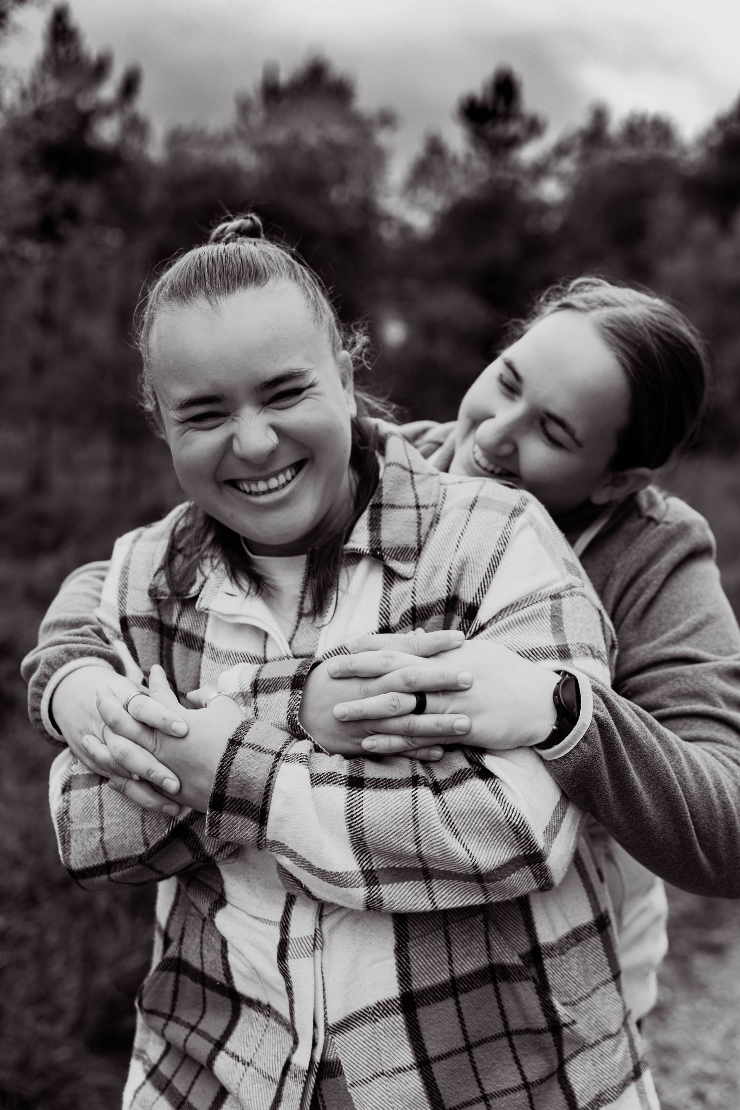 Two women laughing playfully as one hugs the other from behind in black and white.