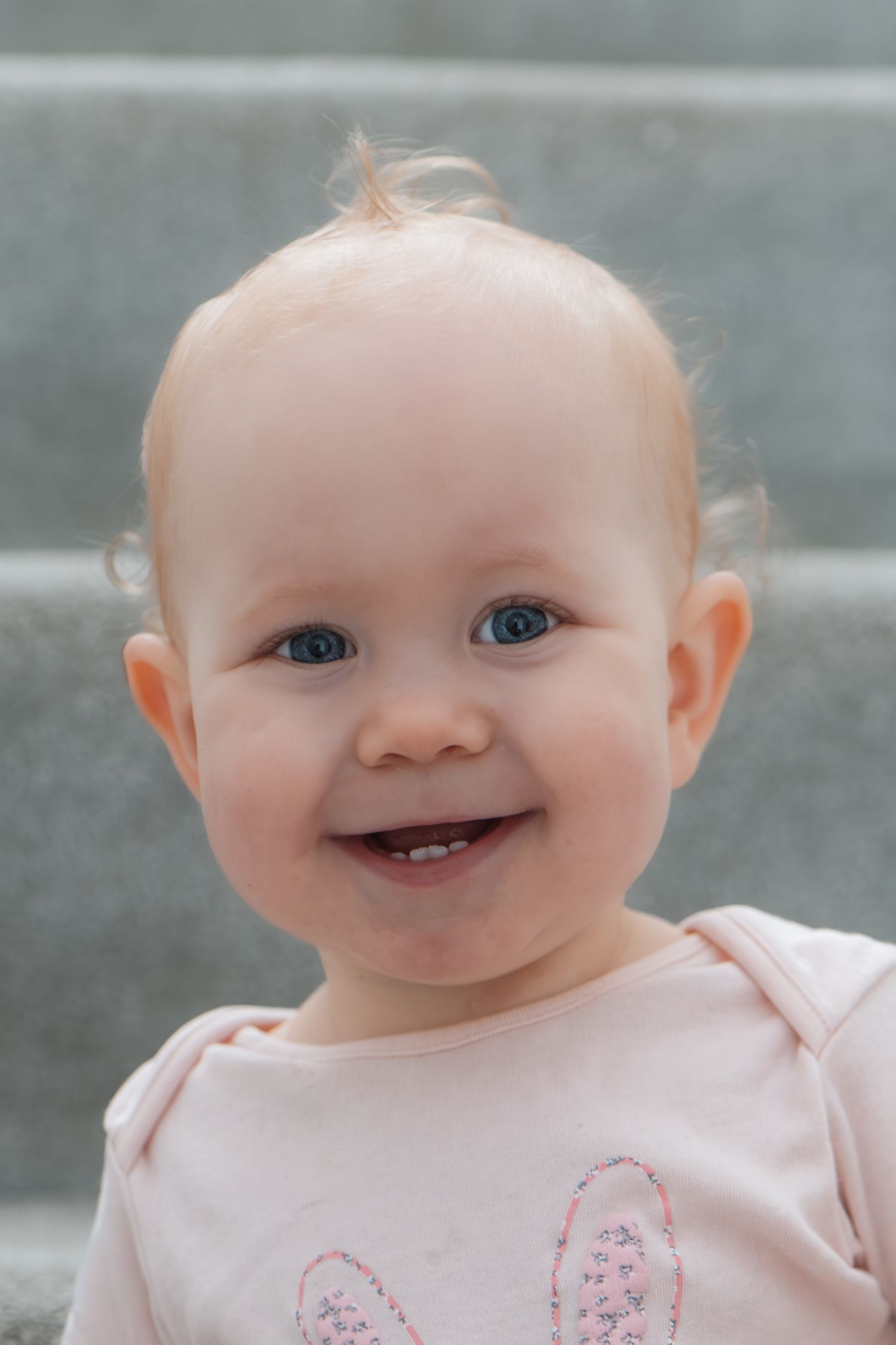 Happy baby girl with blue eyes and two bottom teeth smiling in a pink bunny onesie.