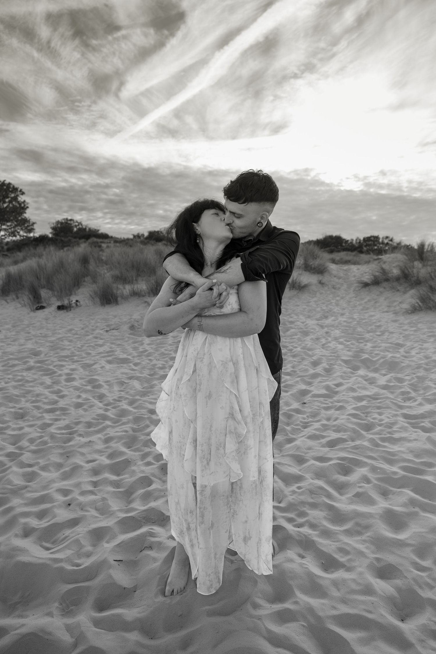 Black and white photo of a couple embracing and kissing on sandy beach dunes under a dramatic cloudy sky.