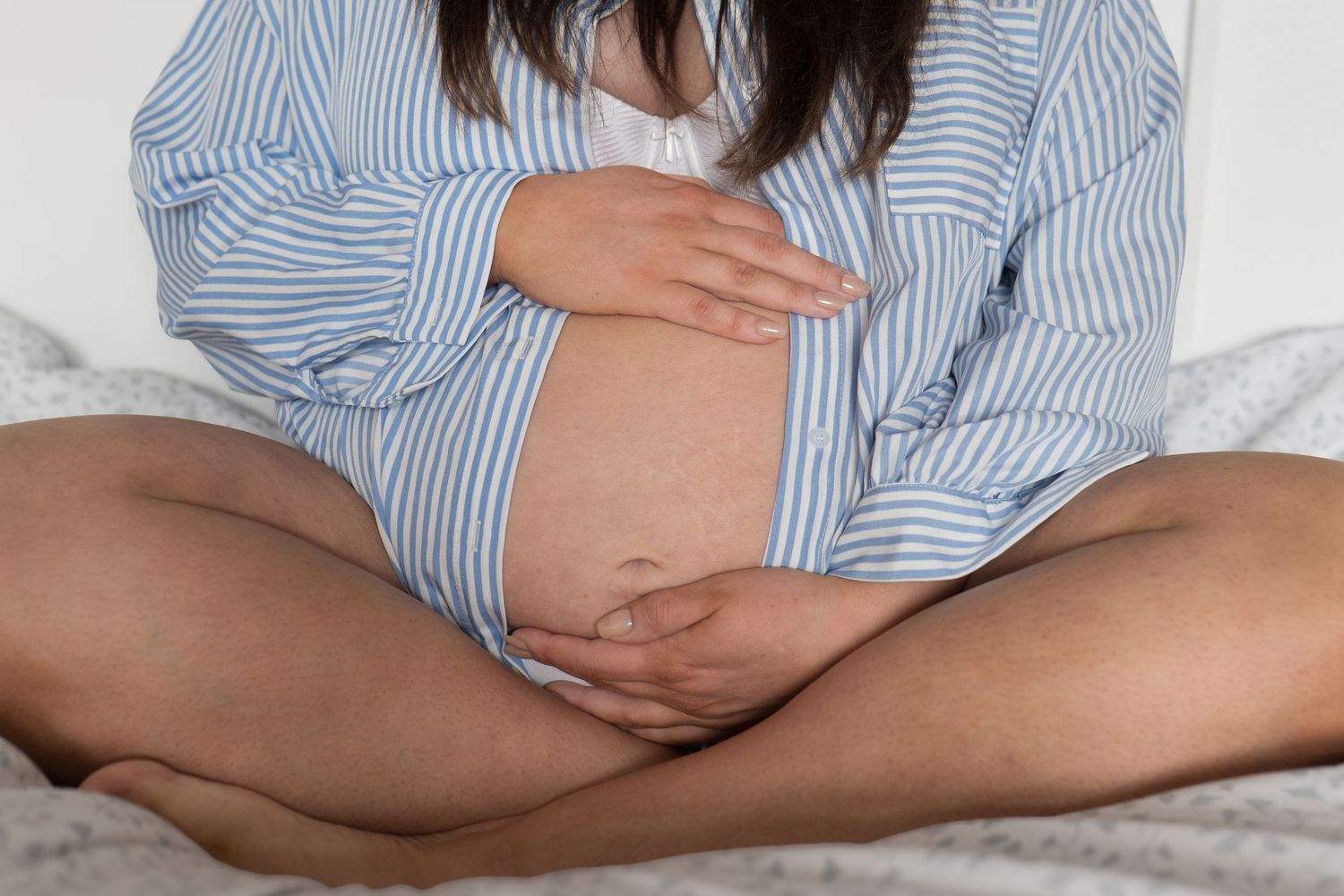Close-up of pregnant woman in striped shirt sitting cross-legged on bed holding her bump.