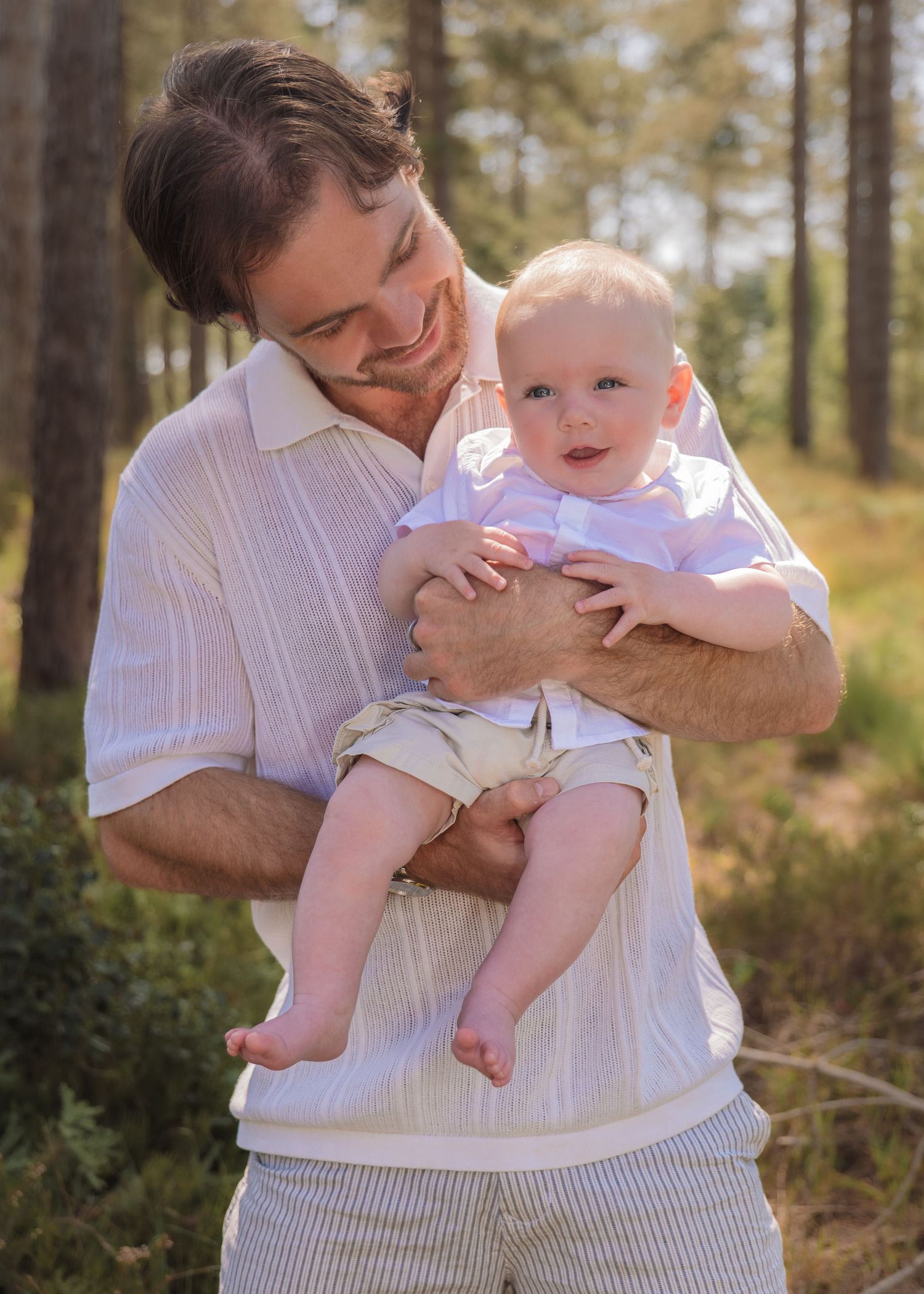 A smiling man holds a happy baby in a sunlit forest setting, both dressed in casual summer outfits.