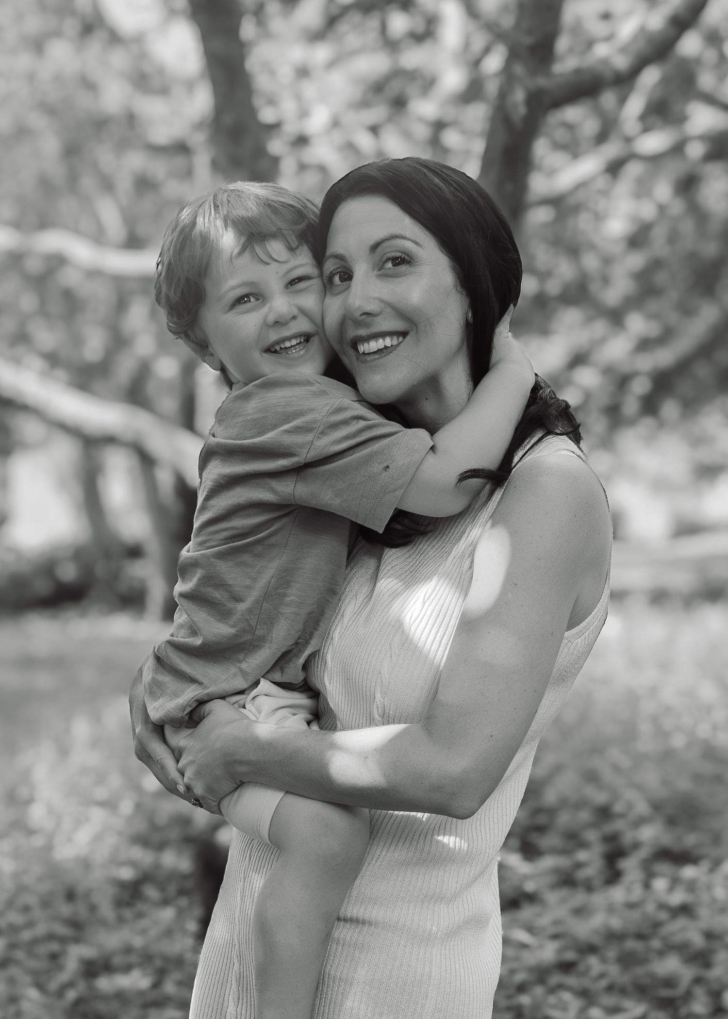 Black and white photo of a smiling woman holding a happy young boy outdoors among trees.