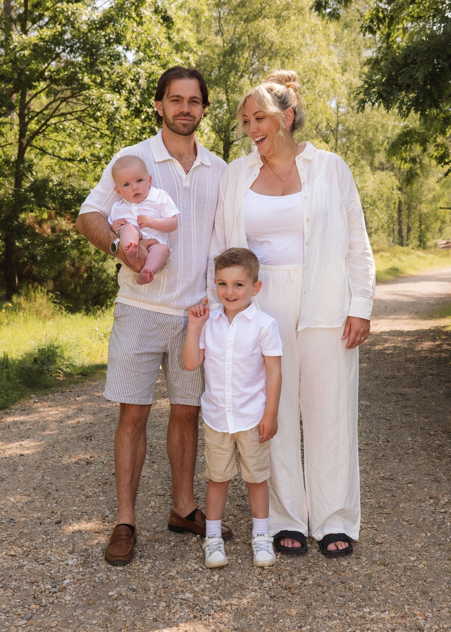 Happy family of four posing on a sunny gravel path, parents with toddler and baby dressed in white and beige.