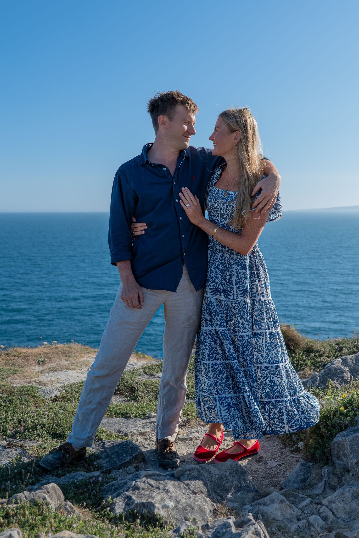 Happy couple embracing on rocky cliff with stunning blue ocean and clear sky behind them.