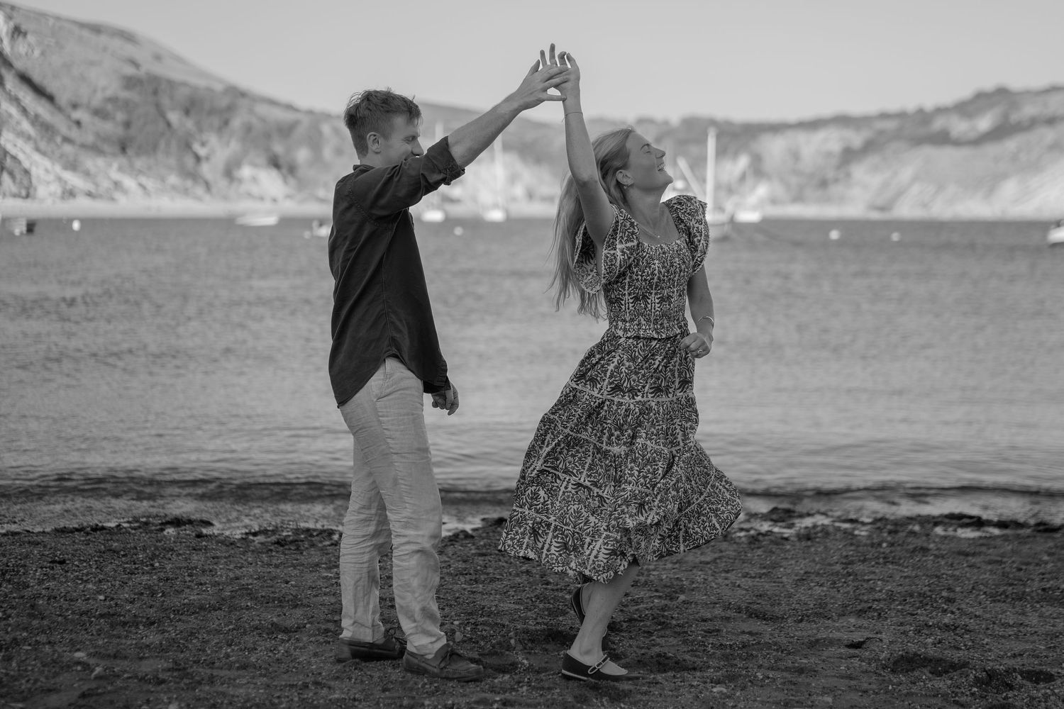 A couple dances together on a rocky beach with ocean and cliffs in the background in this black and white photo.