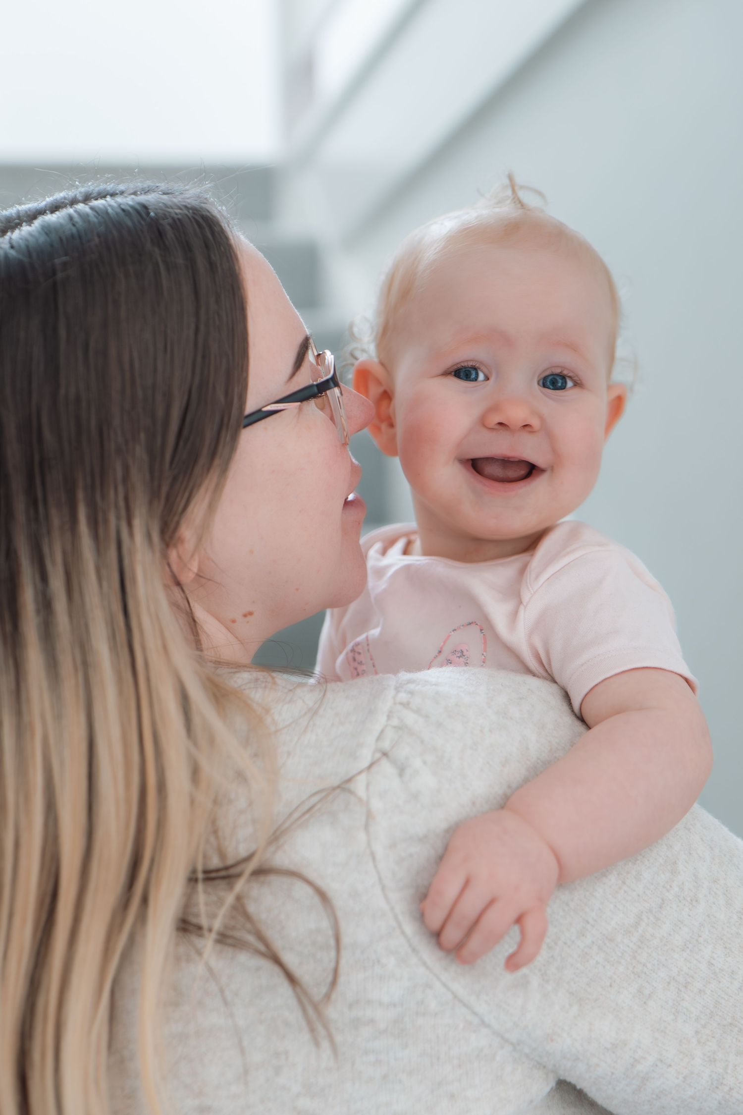 Mother with glasses holding her laughing blue-eyed baby girl in a bright indoor setting.