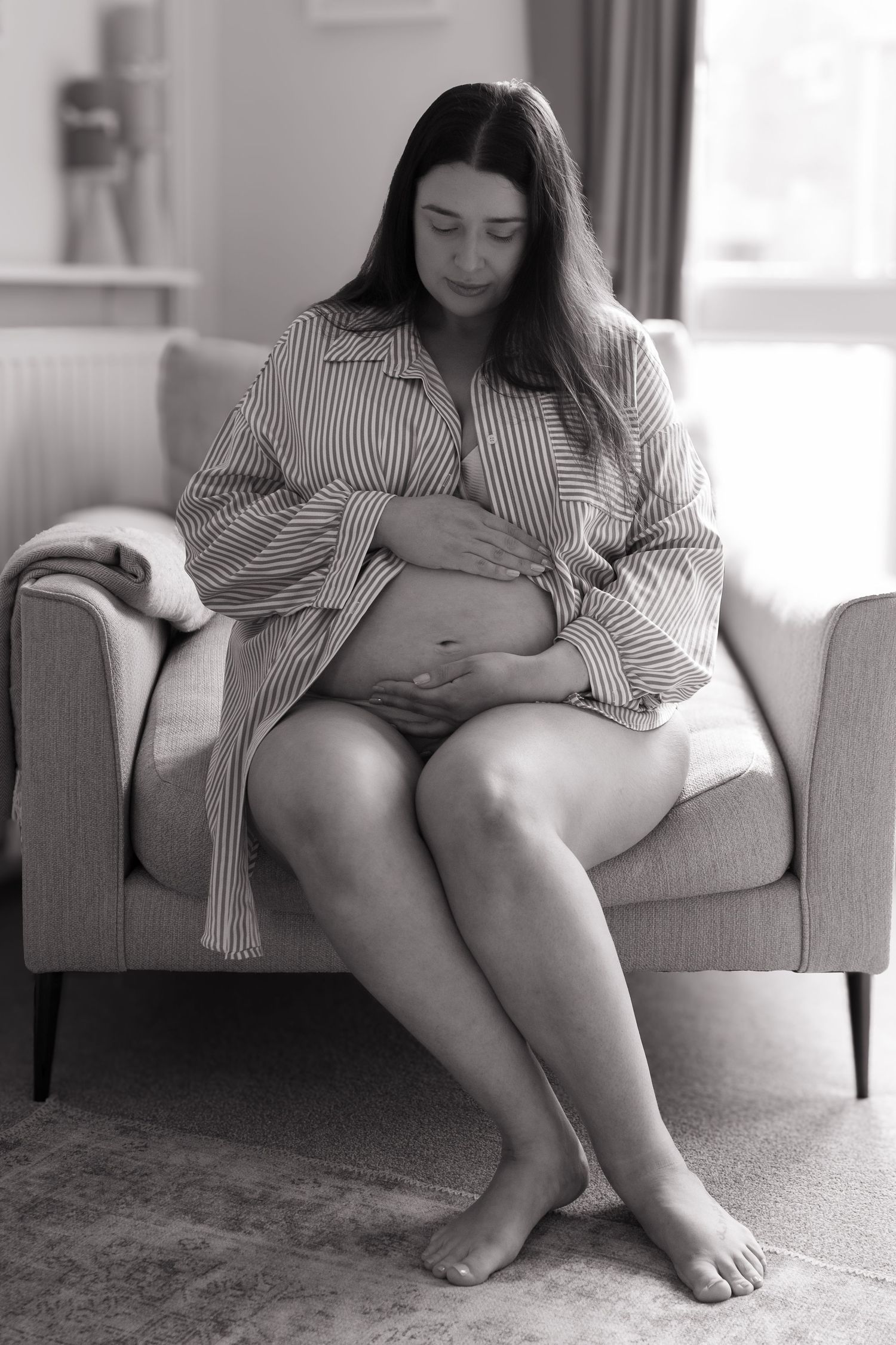Black and white full body photo of pregnant woman in striped shirt sitting in armchair.