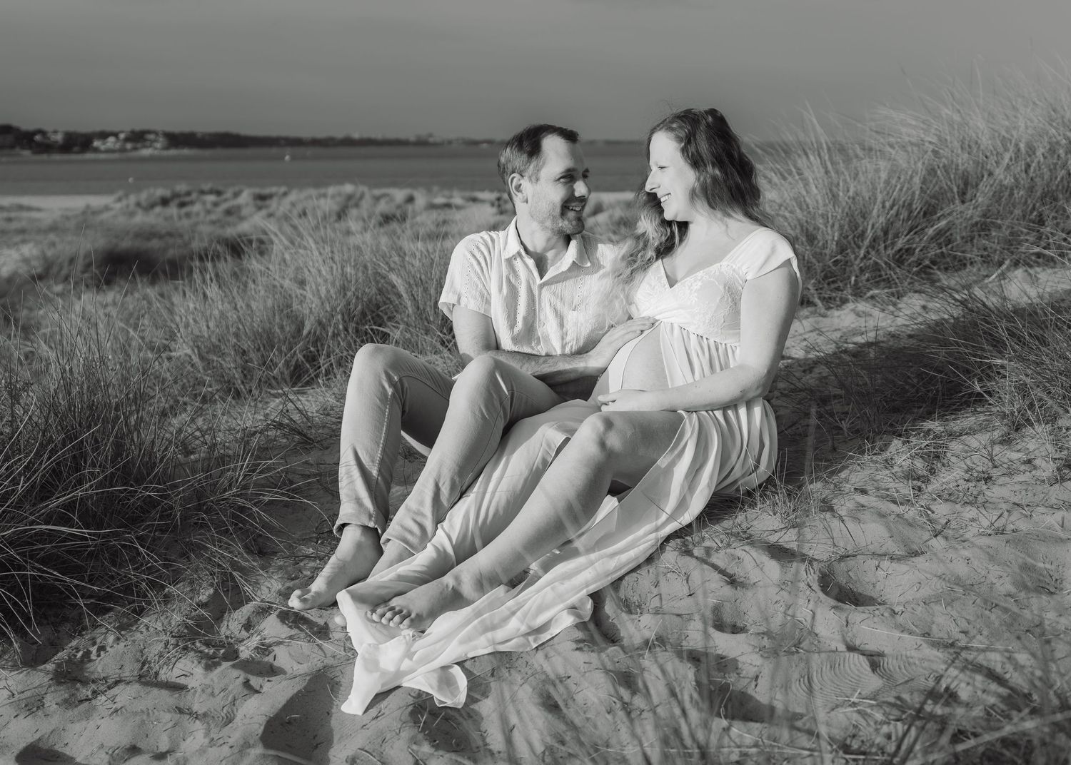Black and white beach maternity photo of a couple sitting on sand dunes, smiling at each other, with ocean in background.