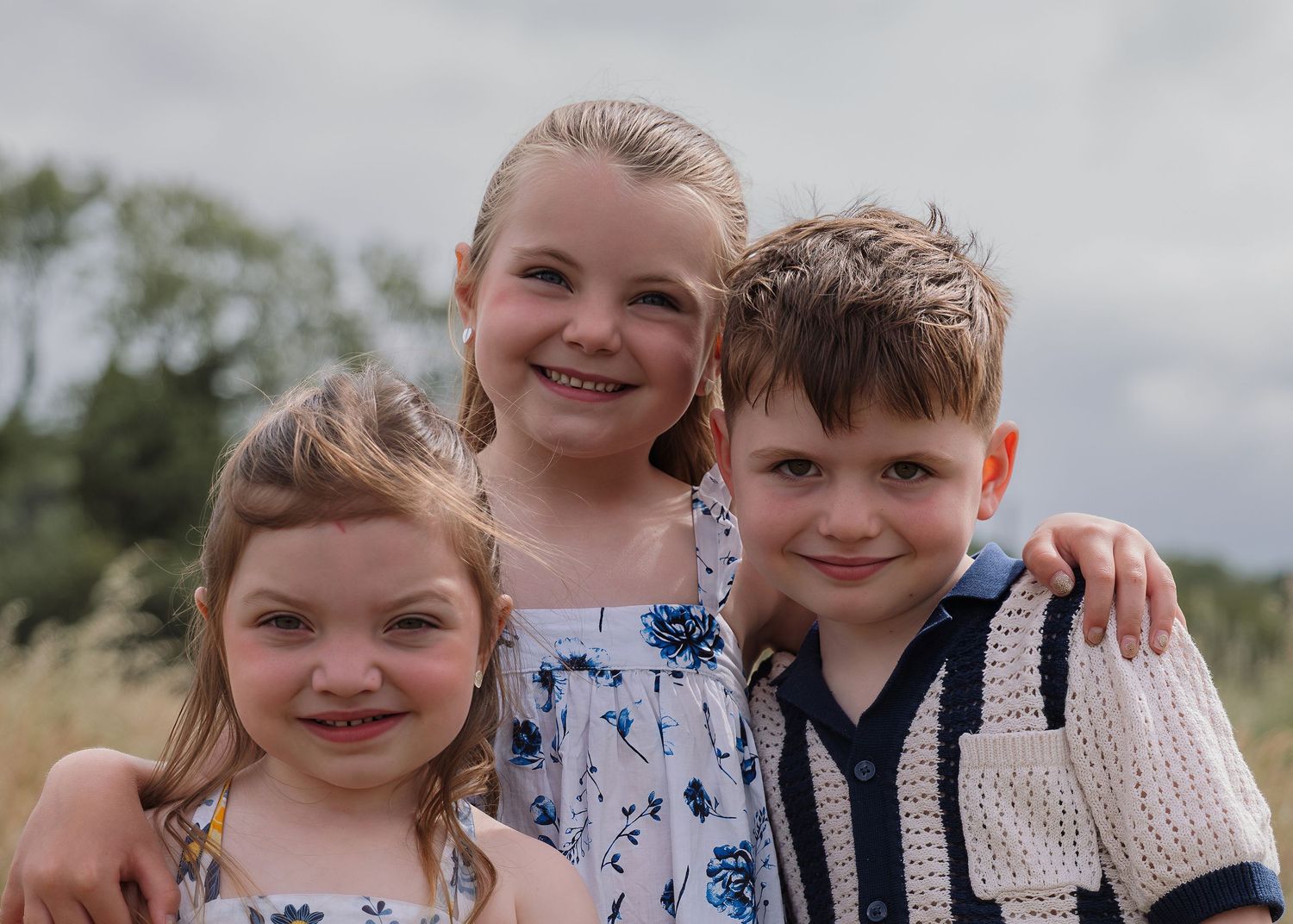 Three smiling children posing outdoors in a field, wearing casual summer clothes on a cloudy day.